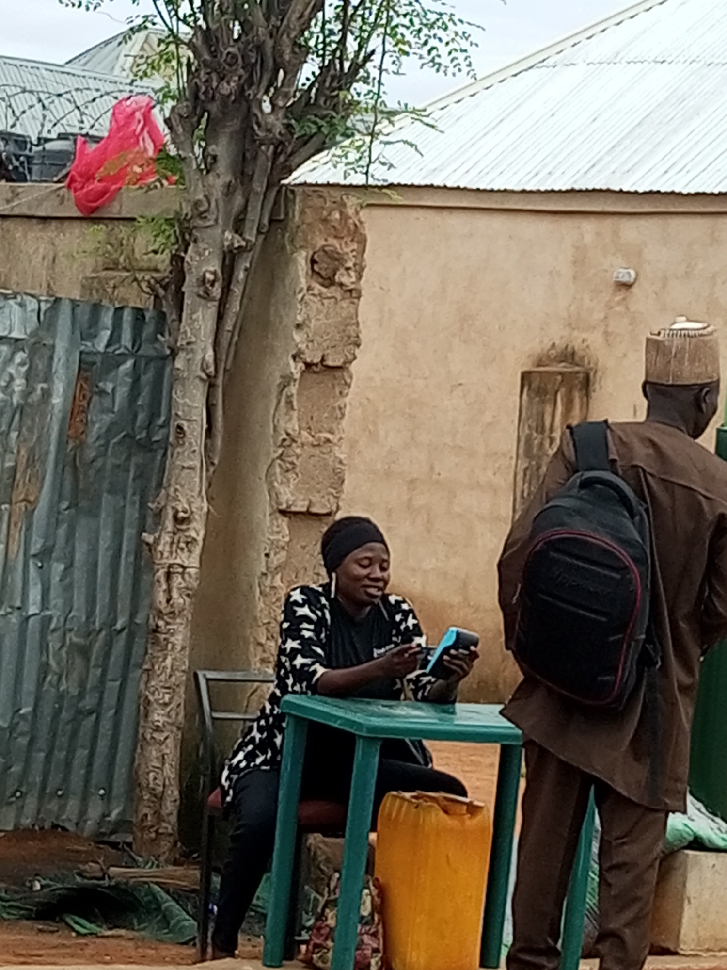 In Tempure, Nigeria, a woman wearing a black headscarf and a black and white patterned shirt sits at a green plastic table. She is holding a blue and black point-of-sale device. Beside the table is a large yellow plastic container. To her right, a man wearing a brown traditional outfit and a cap stands facing away from the camera. He has a black backpack slung over his shoulders. Behind them, a rough-textured building with a corrugated metal roof is visible. A tree with a gnarled trunk stands to the left of the woman, and a section of corrugated metal sheeting is also present. A bright red fabric is draped over a wall to the left of the tree. The ground appears to be dirt.