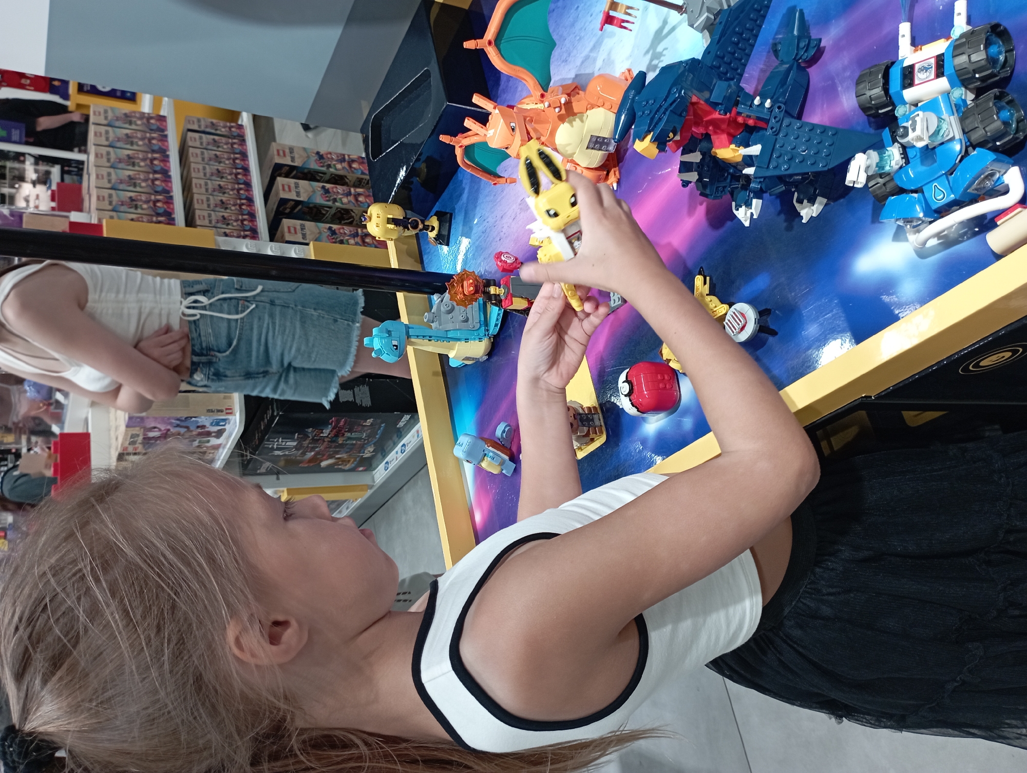 The image shows a young girl playing with LEGOs at a table in what appears to be a store. The girl is wearing a white tank top with black trim and a black tulle skirt. She is holding a yellow LEGO Pokemon in her hands, and there are other LEGO Pokemon figures and vehicles on the table. The table's surface is a printed mat with a galaxy design. In the background, there are shelves stocked with LEGO boxes, indicating the setting is likely a toy store or a dedicated LEGO display area. The presence of other shoppers and retail shelving suggests an indoor commercial environment. The specific location in Katowice, Poland, cannot be determined from the image alone, as there are no identifying landmarks or signage visible.
