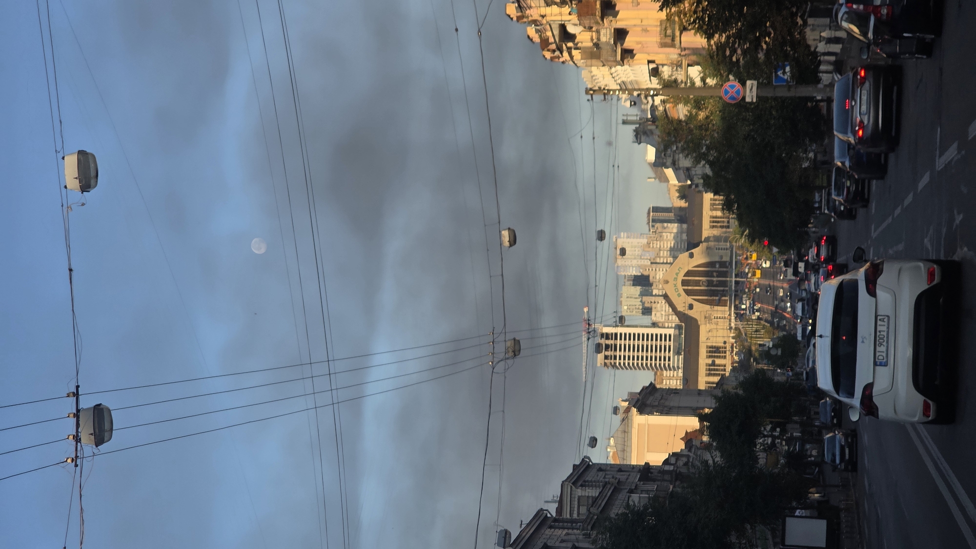 The image captures a street scene in Kyiv, Ukraine, facing a large building with "ВОКЗАЛ" (Station) written across its facade, suggesting a railway station. The perspective is from a vehicle, looking down a tree-lined avenue filled with traffic, including cars and buses. Overhead, a network of wires and what appear to be suspended cable car cabins crisscross the sky. The sky is partly cloudy, with a visible moon. Buildings line both sides of the street, exhibiting varying architectural styles, some appearing older and ornate, others modern and tall. The overall impression is of a bustling urban environment with a mix of transportation and infrastructure.