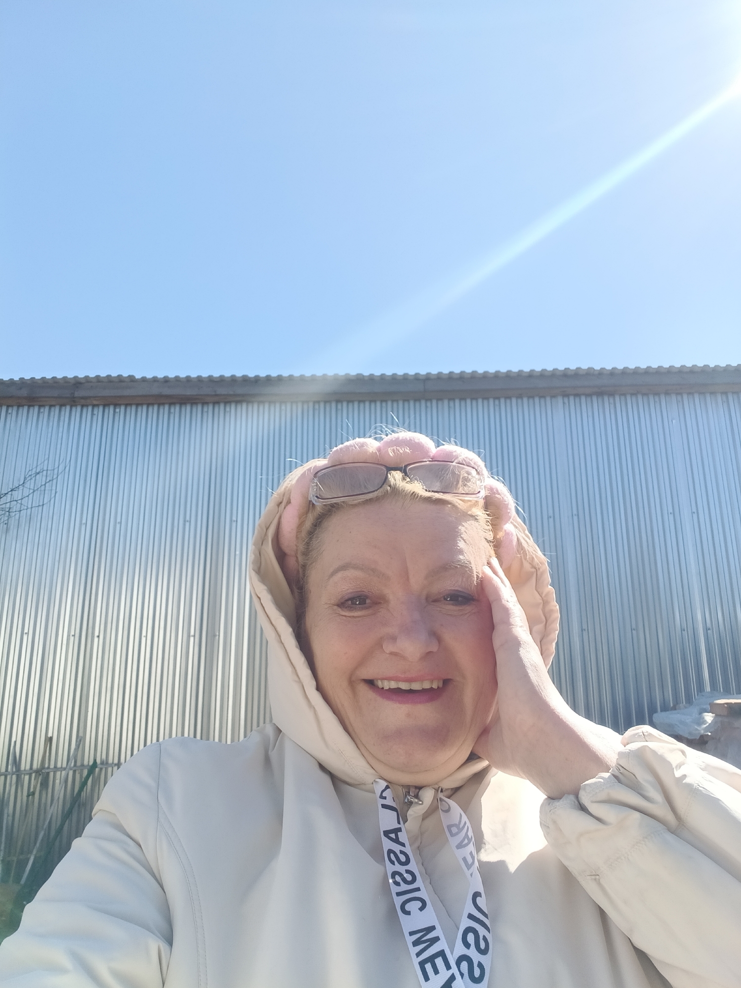 The image shows a middle-aged woman smiling at the camera. She is wearing a light-colored hooded jacket and sunglasses on top of her head. She is touching her face with her hand. The background consists of a corrugated metal wall and a clear blue sky with a sun glare. The location is Красный Маяк, Russia.