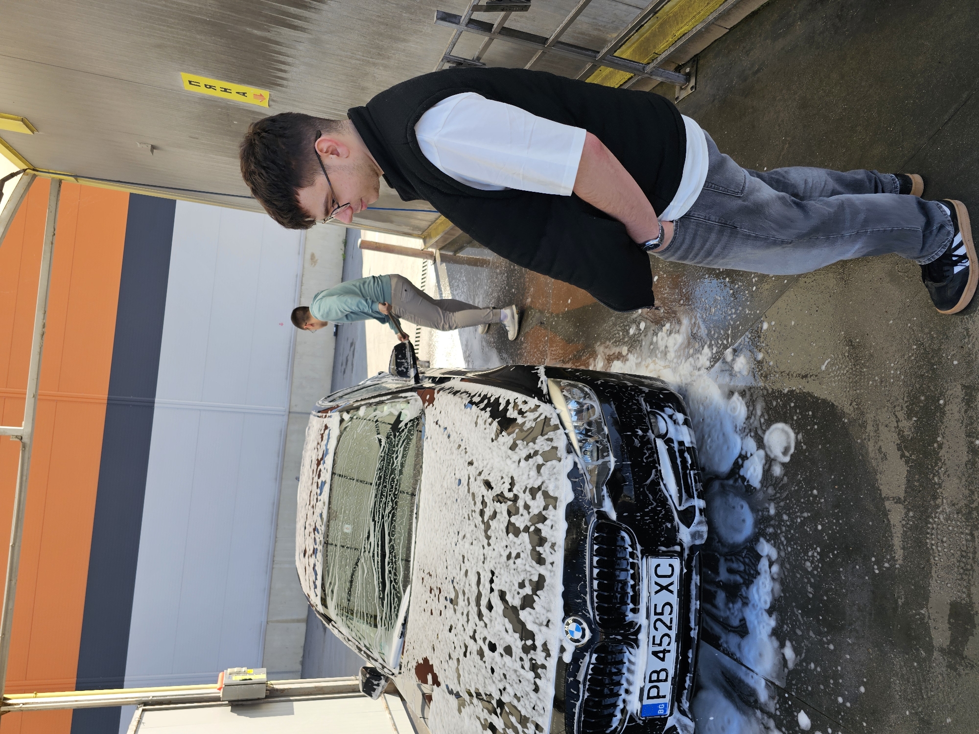 The photograph shows two people washing a black BMW car at a self-service car wash. The car, covered in soap suds, has a license plate reading "PB 4525 XC." The first person, in the foreground, is an adult male wearing glasses, a white t-shirt, a black vest, gray jeans, and black sneakers. He is standing with his hands in his pockets, looking at the car. The second person, in the background, is also an adult male wearing a light green shirt and gray pants, bending down and holding a pressure washer wand. The car wash location appears to be outdoors with orange, gray, and white wall panels. The location is likely in Plovdiv, Bulgaria, based on the license plate information.
