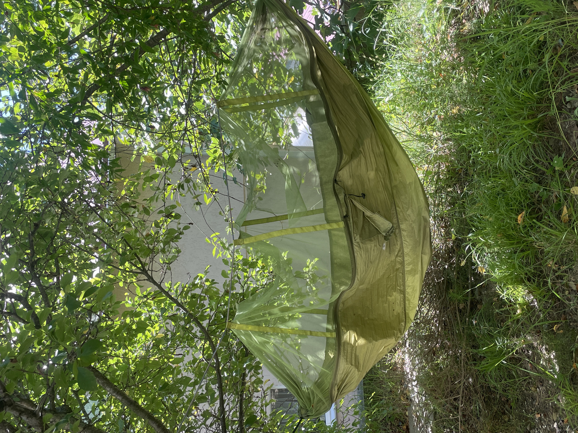 An olive green hammock with a built-in mosquito net is suspended between two tree branches in an outdoor setting. The hammock is made of a durable fabric and features a fine mesh net that encloses the sleeping area. The net is supported by several vertical straps that attach to the hammock's fabric. Sunlight filters through the lush green leaves of the trees, casting dappled shadows on the hammock and the surrounding grassy ground. In the background, a light-colored building with windows is partially visible through the foliage. The overall scene suggests a peaceful and relaxing outdoor environment.