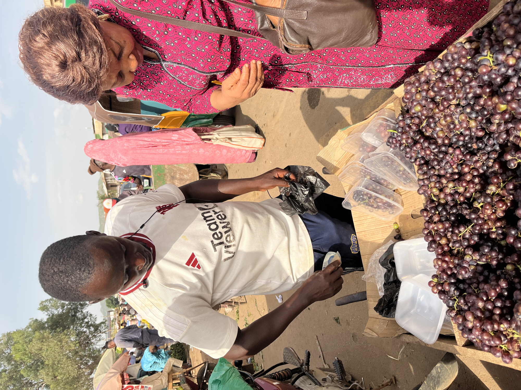 At a street market in Rafin Par, Nigeria, a Black man wearing a white t-shirt with "Team Viewer" printed on it and dark blue shorts is selling a large pile of dark purple grapes. He holds a few bills in his right hand, appearing to be completing a transaction with a Black woman. The woman, wearing a pink patterned dress and a brown shoulder bag, looks down towards the grapes and her hands. The grapes are displayed in a wooden crate, with some already packaged in clear plastic containers. In the background, other people and market stalls are visible, as well as a tricycle commonly used for transportation in the region. The ground is dusty, typical of an outdoor market.