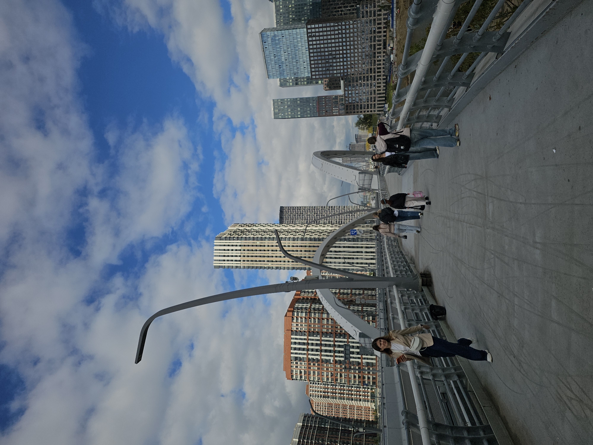 The image shows a pedestrian bridge with several people walking on it. In the background, there are several modern buildings, some with glass facades, and a tall skyscraper. The sky is blue with scattered white clouds.
More specifically, the scene depicts the pedestrian bridge over the Moscow Canal, located near the Moscow City business district. The buildings in the background are characteristic of the modern architecture found in this part of Moscow, including residential complexes and office towers.
In the foreground, a woman in a beige jacket and dark pants is walking towards the viewer, holding a smartphone. Further along the bridge, a group of people are walking away from the camera. One person in the group, wearing a backpack, appears to be taking a picture or looking at something with their phone.
The bridge itself is made of concrete and metal, with railings on either side. The architectural style of the bridge, with its arched supports, is a distinctive feature. The overall impression is of a contemporary urban landscape on a clear day.