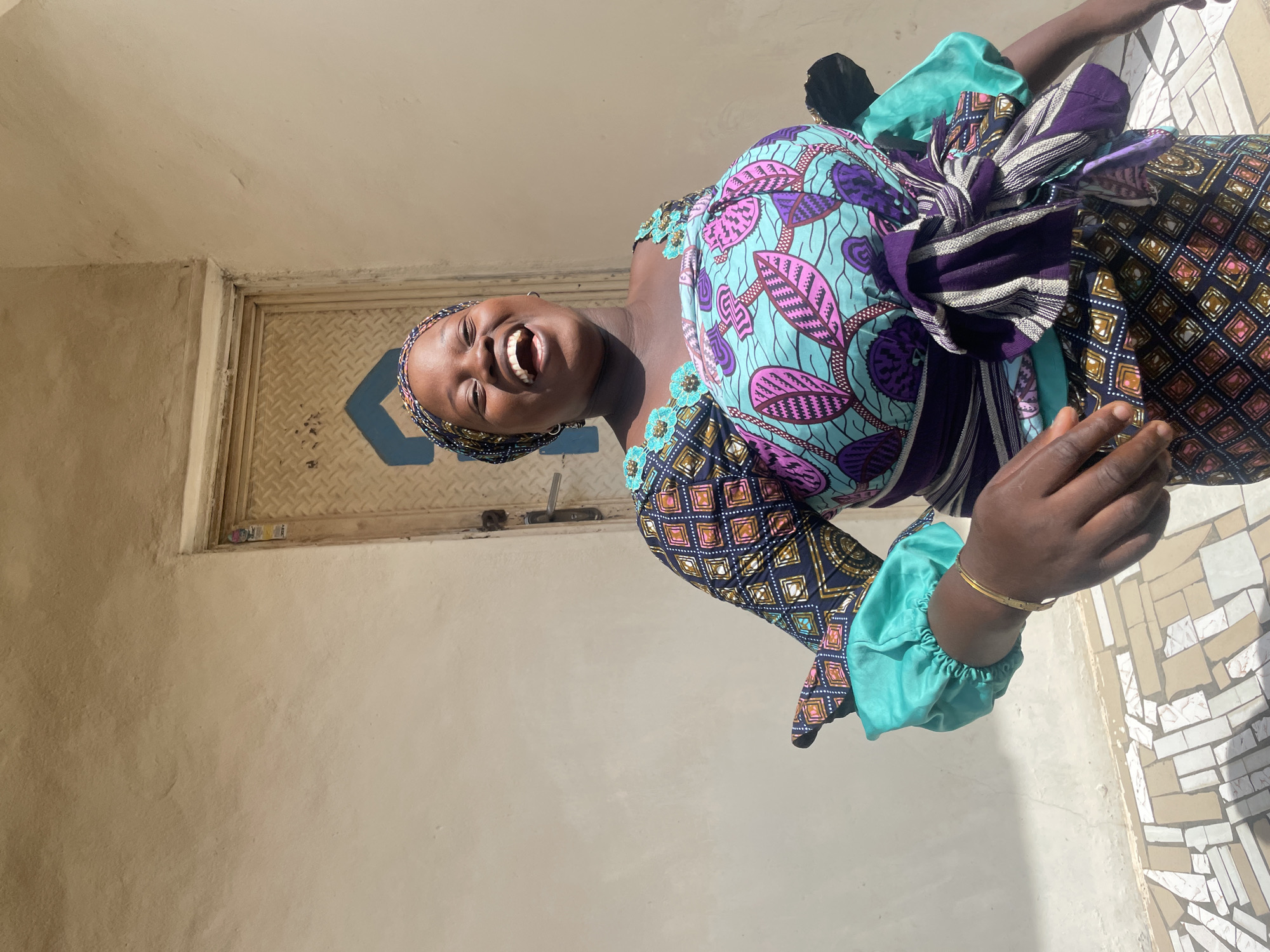 A woman in Lokoja, Nigeria, stands smiling, her head tilted back in apparent joy. She is wearing a colorful, patterned dress in shades of purple, blue, and green, with a matching head wrap. A striped purple and white sash is tied around her waist. The background shows a beige-colored wall with a metal door featuring a blue, geometric design. The flooring has a light-colored, patterned tile design. The photograph appears to be taken outdoors in bright sunlight.