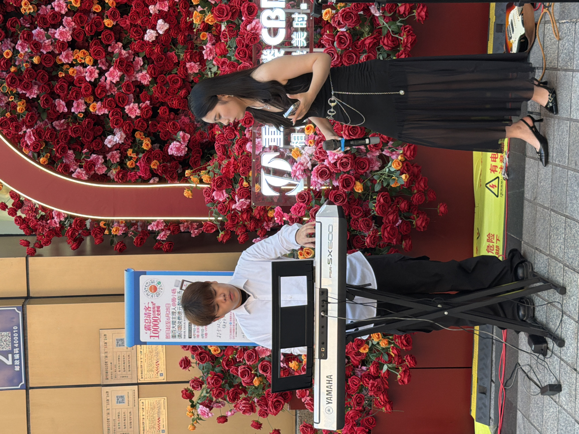 A man in a white shirt and black pants plays a Yamaha PSR-SX600 keyboard in front of a large floral arrangement of red and pink roses. To his right, a woman in a black dress holds a microphone and a mobile phone. Both are positioned outdoors on a paved surface with some yellow caution tape visible. A guitar and cables are also present. The background features a building facade and signs, one of which indicates "Yuzhong District, China".