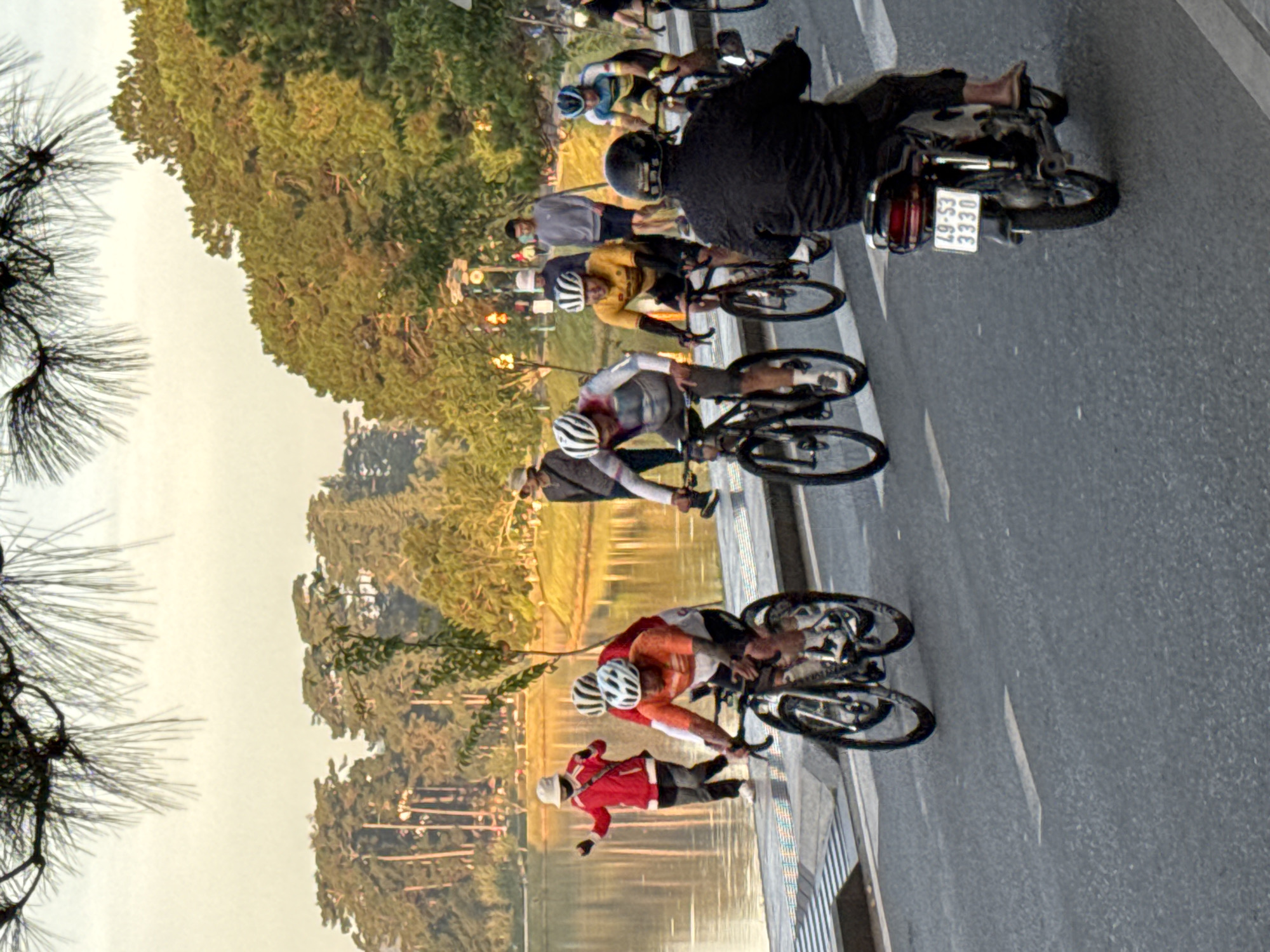 The image depicts a scene in Da Lat, Vietnam, likely near Xuan Huong Lake, a popular recreational area. Several cyclists are riding on a paved road alongside the lake. In the foreground, a person on a motorcycle, wearing a dark jacket and pants, is visible from the back, with a license plate reading "49-S3 3330". Behind them, multiple cyclists are in motion. One cyclist in an orange and white jersey is wearing a helmet. Another cyclist in yellow and black attire, also wearing a helmet, is riding a bicycle. Further in the background, a person in a red jacket and a white hat appears to be standing by the water's edge, possibly interacting with someone off-camera. Other individuals are seen walking or cycling in the middle ground, and lush green trees and foliage line the background, suggesting a park-like setting. The lighting indicates it's likely morning or late afternoon, with soft sunlight illuminating the scene.