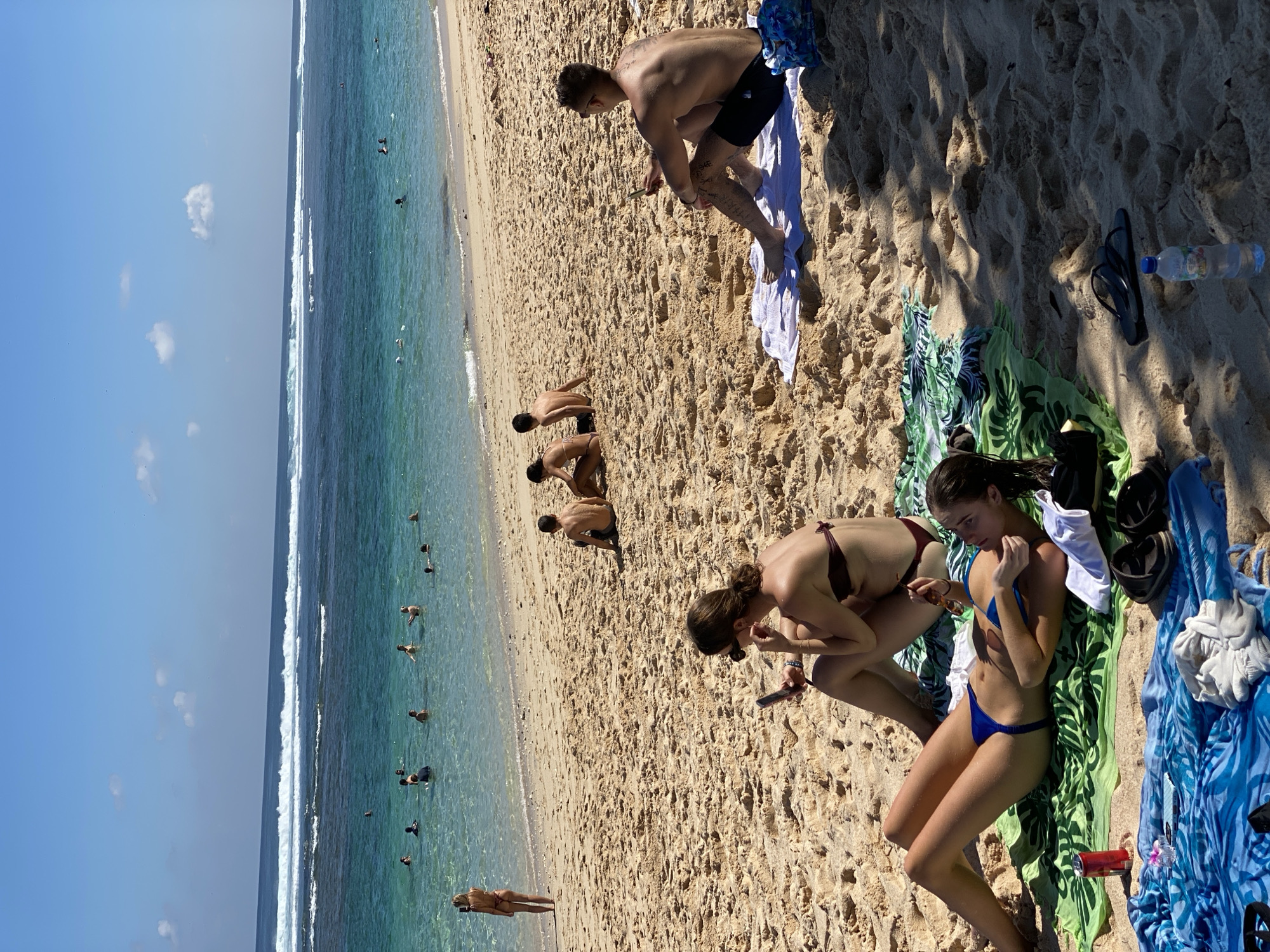 The image depicts a sunny day at a beach in Kuta Selatan, Indonesia. The foreground is dominated by sand, with several people relaxing on towels and the sand itself. In the center foreground, a young woman in a blue bikini lies on her back on a green towel, holding sunglasses. Next to her, another woman in a maroon bikini sits with her legs crossed, looking at her phone. Further back, a man with tattoos on his arms and legs sits on a white towel, also looking at his phone. Behind them, two men are sitting in the sand. In the midground, several people are visible walking and wading in the shallow turquoise water. The background shows a clear blue sky with a few white clouds and the ocean with white-capped waves. The overall scene conveys a relaxed atmosphere of people enjoying a day at the beach.