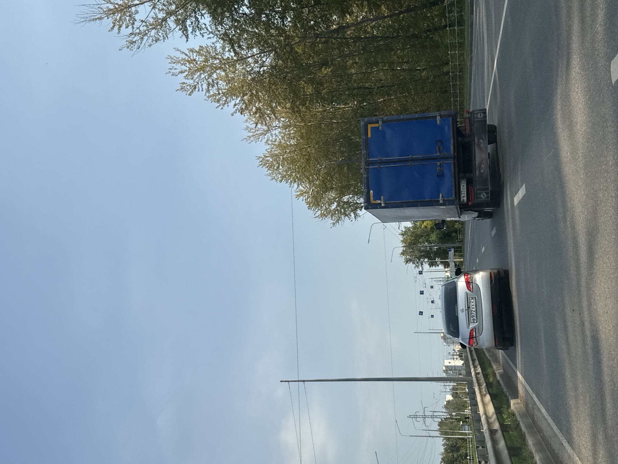 The image shows a view from a vehicle traveling on a highway. In the foreground, a silver car is visible on the left, and a blue truck is on the right. The truck has an image of eyes on its rear bumper. Trees line the right side of the highway, and power lines and streetlights are visible in the distance. The sky is blue with a few wispy clouds. The location is a road in an urban or suburban area, likely in Russia, given the license plates on the vehicles.