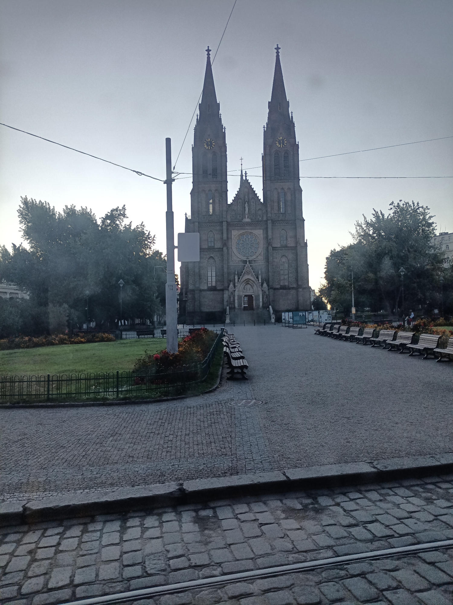 The image shows a view of the Church of Saint Peter and Paul in Vyšehrad, Prague, Czechia. The church features two prominent spires with clocks and a large rose window above the main entrance. The foreground is a cobblestone plaza with rows of empty wooden benches. To the left, there is a small park area with a green lawn, flower beds, and a black metal fence. Trees line the sides of the plaza and park. Overhead, power lines and a utility pole are visible. The sky is overcast and a pale gray. There are no visible people actively engaged in any interactions, though a few distant figures can be seen in the park to the right. The scene appears to be an outdoor public space in a European city, likely during the daytime based on the ambient light, despite the cloudy conditions.