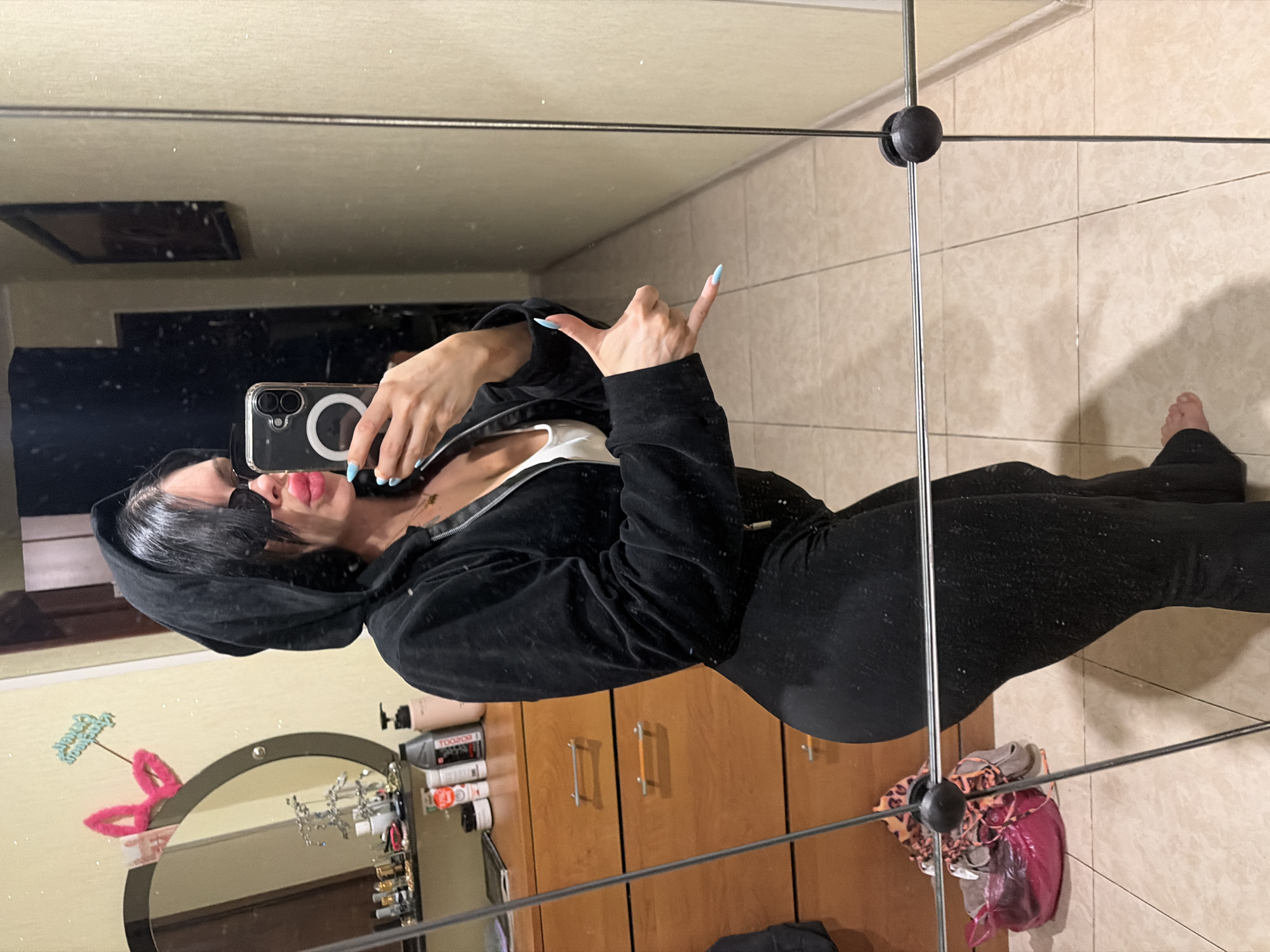 A woman is taking a mirror selfie in a dimly lit room. She is wearing a black hooded jacket, a white tank top, and black pants. She has sunglasses on and is making a shaka sign with her right hand. In the background, there is a dresser with various items on it, a mirror, and a framed picture on the wall. The floor is tiled. This image was not taken in Ufa, Russia.
