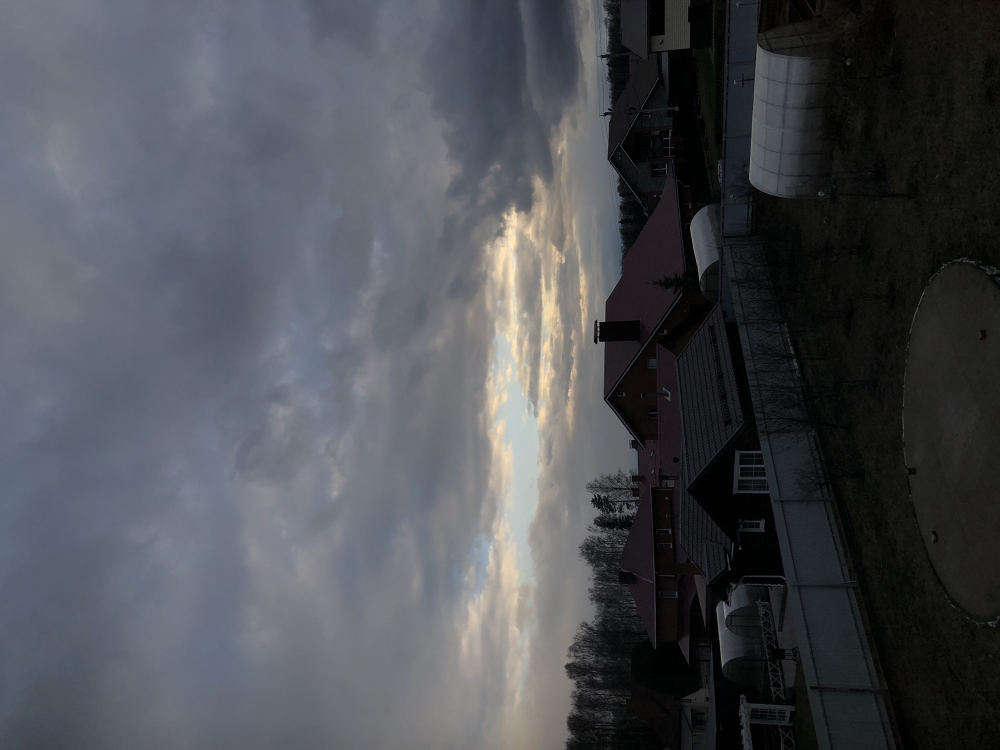 The image depicts a residential area under a dramatic, cloudy sky. When viewed in standard orientation, the sky occupies the upper portion, filled predominantly with heavy, dark grey clouds. A prominent vertical band of brighter, lighter clouds with hints of blue sky cuts through the darkness, suggesting the sun is either breaking through or at a low angle (setting or rising).
Below the sky, several houses with pitched roofs are visible. The roofs are in varying shades of dark red-brown and dark grey/black. Some houses show dark-colored walls, with one featuring white window frames that stand out. Multiple chimneys can be seen on the rooftops. A line of bare, deciduous trees stretches across the horizon, indicating a colder season like late autumn or winter.
The ground areas visible are dark, likely covered with grass or bare earth. In the middle and foreground, fences delineate property lines. On the right side of the image, two tunnel-shaped greenhouses with translucent white coverings are situated on the ground. A large, flat, circular structure, possibly a pool cover or a trampoline, is partially visible in the bottom right corner.
The overall lighting suggests either a late afternoon/early evening or early morning scene, with the diffused light filtering through the clouds. The mood is subdued due to the heavy cloud cover, but the bright band of sky introduces a contrasting element of light. There are no people or explicit activities taking place, and no visible text can be identified. The setting appears to be a quiet suburban or rural residential area.