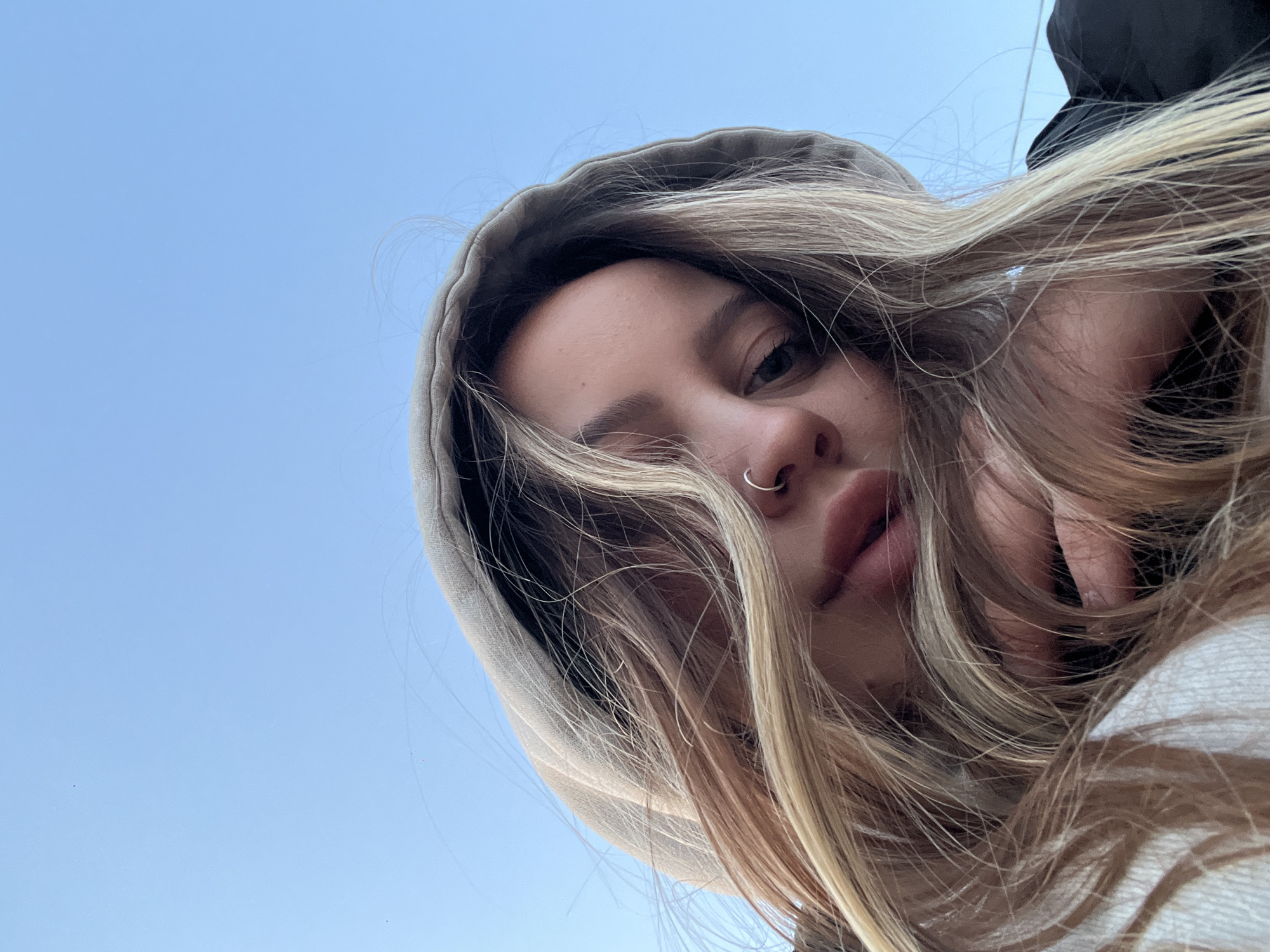 A low-angle close-up captures a young woman looking down towards the camera against a clear, light blue sky. Her face is centered, framed by long, wavy blonde and light brown hair that cascades around her. She wears a light-colored hooded garment, possibly a hoodie, with the hood pulled up, partially obscuring the top and sides of her head. A silver hoop nose ring is visible on her left nostril. Her eyes are directed towards the viewer with a steady gaze, and her lips are slightly parted. The overall perspective suggests the photo was taken from below, possibly a selfie, with the vast expanse of sky forming the sole background. A dark fabric, possibly part of another garment or an object, is visible in the upper right corner behind her hair. No text is visible in the image. The setting appears to be outdoors during clear daylight in Novopokrovskaya, Russia.