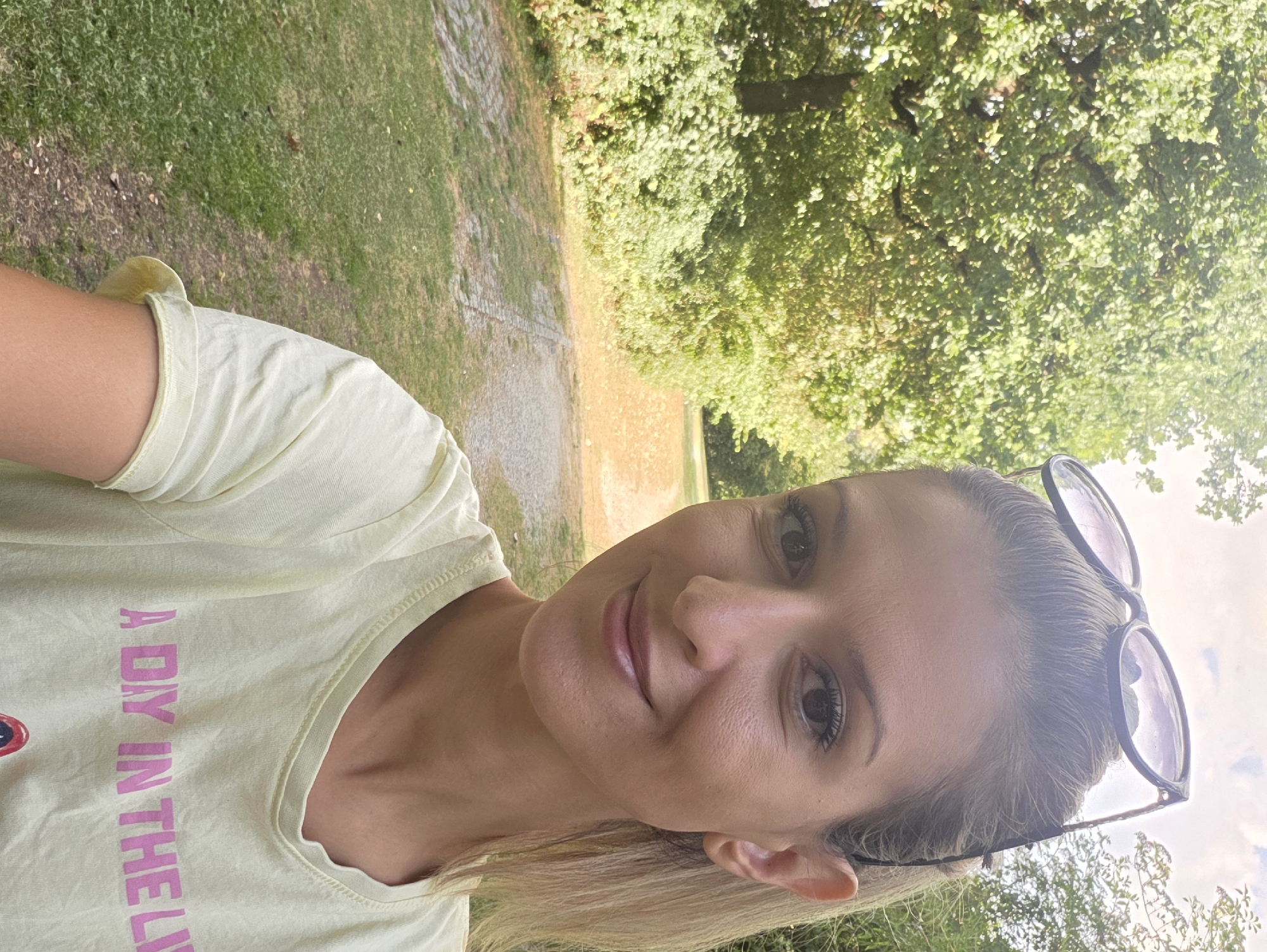 A woman with blonde hair pulled back and sunglasses perched on her head takes a selfie in a park. She is wearing a light yellow t-shirt with pink lettering that reads "A DIY IN THE LI". The background shows a path winding through a wooded area with green trees and grass. The lighting suggests it is daytime. The location is an unidentified park in Munich, Germany.