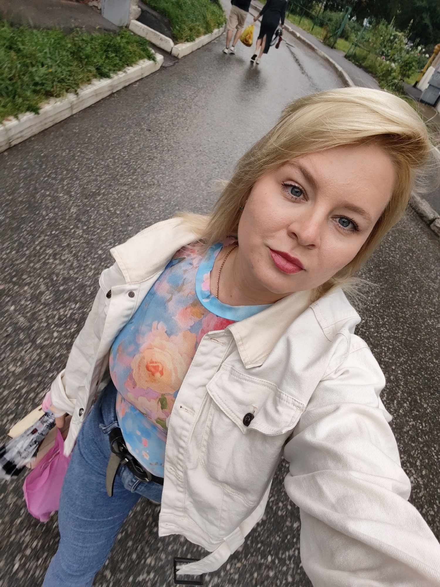 A woman with blonde hair and blue eyes is taking a selfie outdoors. She is wearing a white denim jacket over a blue and pink floral print shirt, and blue jeans. In her left hand, she holds a pink bag and a folded umbrella or fan. She is standing on a wet asphalt surface, likely a sidewalk or street, with a grassy area and a concrete curb to her left. In the background, two people are walking away from the camera, one carrying a yellow bag. The overall environment suggests an urban setting, possibly a park or residential area. The image appears to have been taken in Izhevsk, Russia, based on the provided context.