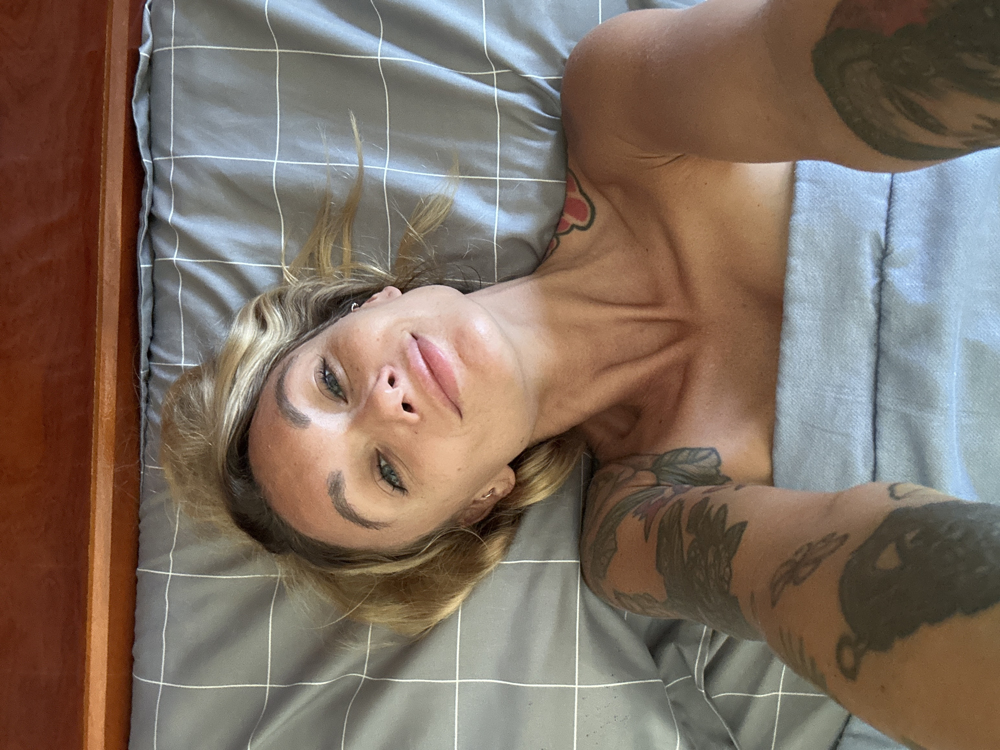 A woman with blonde hair and tattoos lies on a bed with grey, checkered sheets. The bed is positioned near a wooden headboard. The woman is looking directly at the camera with a slight smile. Her body is partially covered by a light blue blanket. The scene appears to be indoors, likely a bedroom, given the bed and headboard. There are no visible elements that would indicate a specific location within the city of Mũi Né, Vietnam.