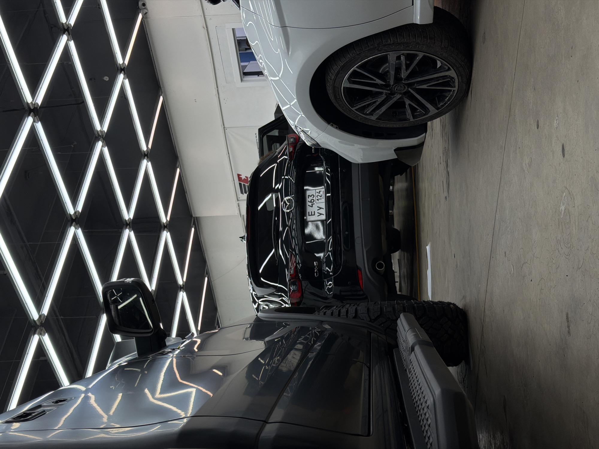 The image captures three cars parked in what appears to be a professional car detailing studio or showroom. The room is brightly lit by an array of white LED tube lights arranged in a prominent, geometric diamond pattern across the ceiling and upper walls, creating striking reflections on the vehicles' highly polished surfaces.
In the foreground, a dark grey or black rugged SUV or truck is visible, featuring large, knobby tires and a black side step. Its glossy paint reflects the studio lights intensely. Behind it, a black Mazda CX-5 SUV is parked, with its rear facing the viewer. The vehicle's license plate, "E 463 YY 124 RUSSIA," is clearly visible, with "124" indicating the Krasnoyarsk Krai region. The model name "CX-5" is also visible on the back. Further in the background, partially obscured by the Mazda, is the front quarter of a white car, possibly another SUV, with a detailed multi-spoke wheel and glossy white paint also reflecting the intense overhead lighting.
The floor is a light-colored concrete surface. A white banner with some red text is partially visible in the background, to the left of the white car. The overall setting suggests a focus on automotive aesthetics, likely showcasing clean, well-maintained, or newly detailed vehicles.