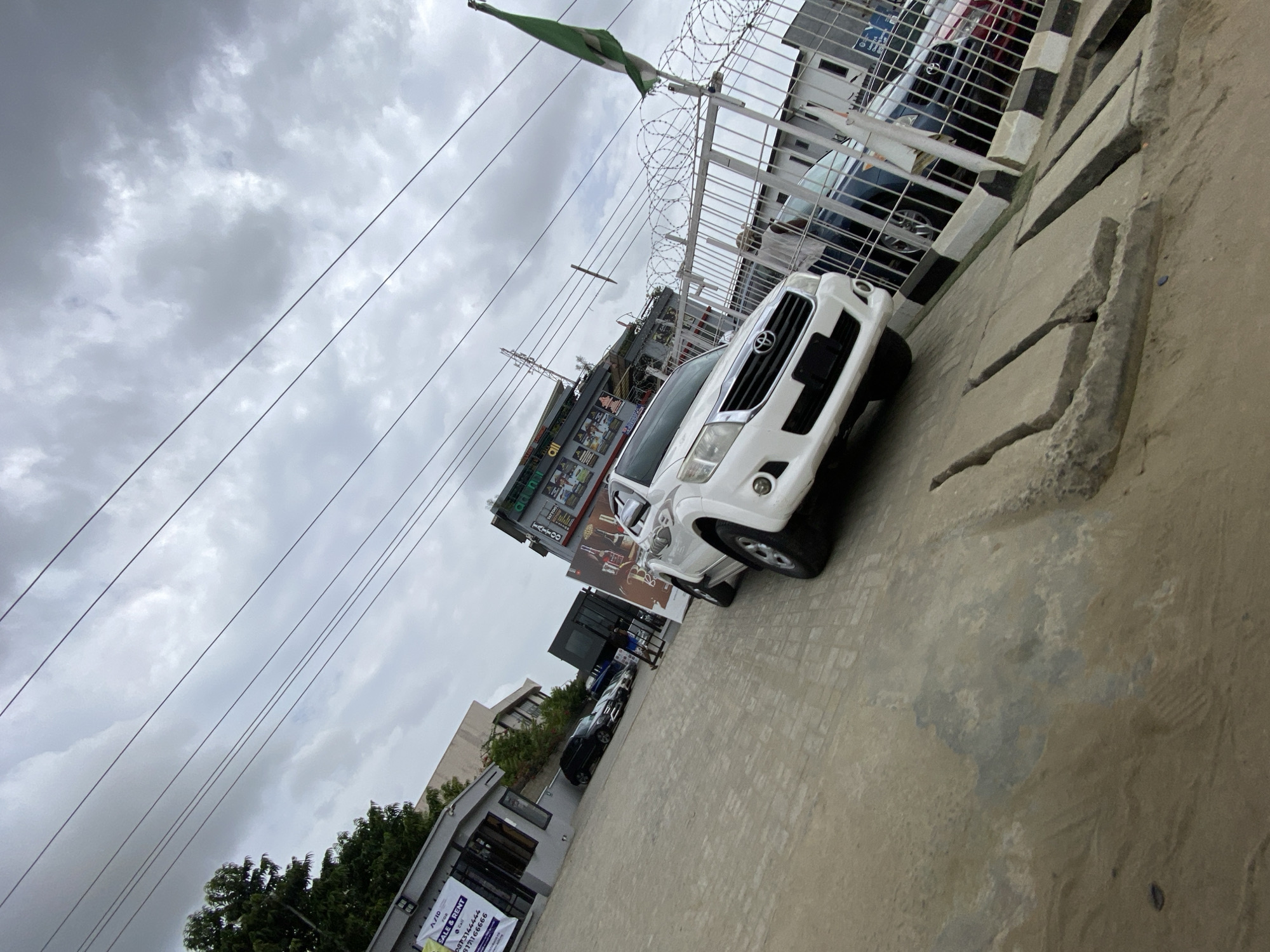 The scene shows a white Toyota Hilux parked on a paved surface in what appears to be an urban setting in Lekki, Nigeria. Overhead are cloudy skies with electrical lines. A green and white flag flies near a fence topped with barbed wire. Buildings are visible in the background, including one with signage. Other vehicles are parked nearby. The pavement has visible cracks and is elevated.