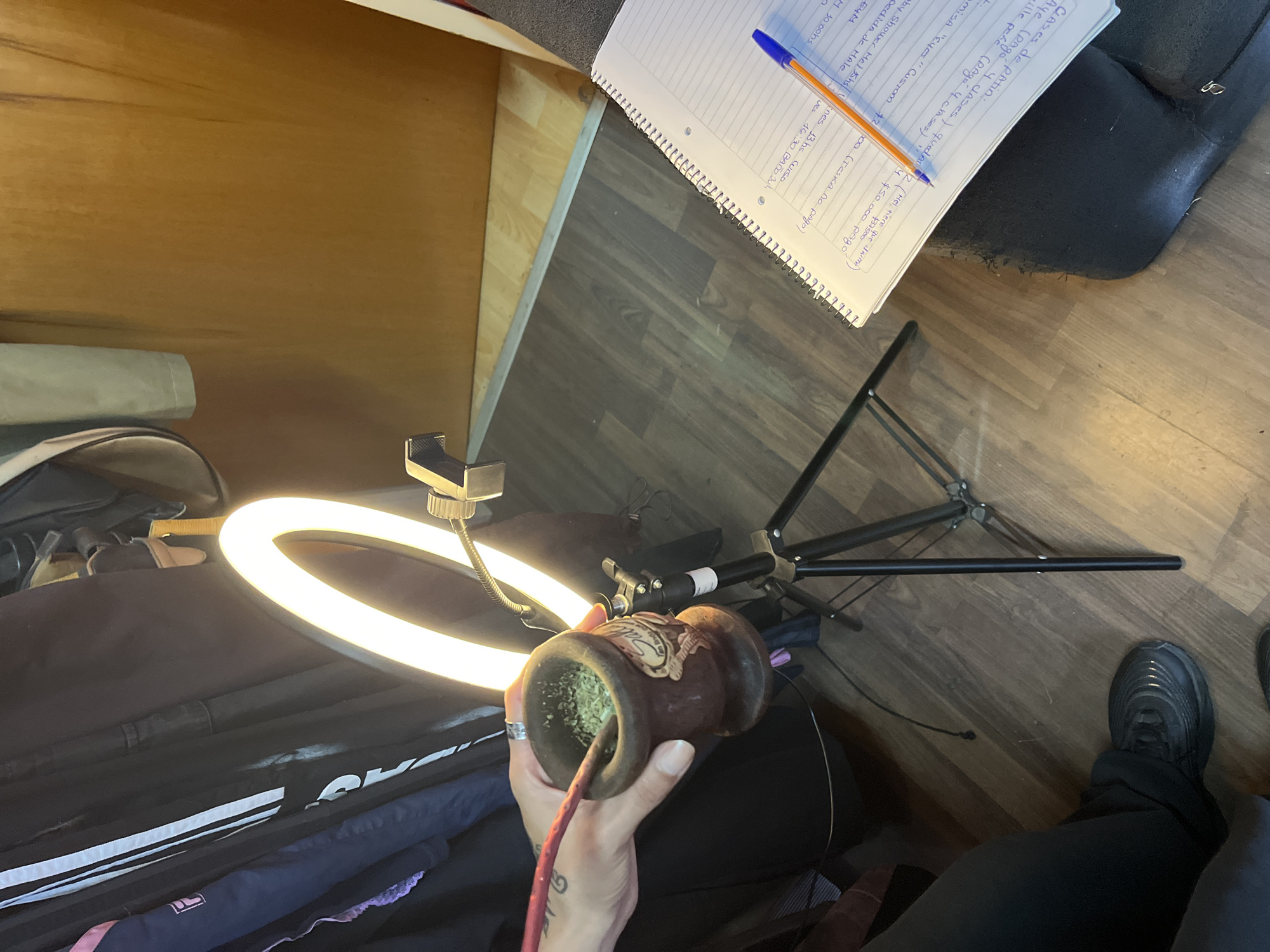 The scene appears to be an indoor setting in Llavallol, Argentina, showing a person preparing what seems to be mate, a traditional South American caffeinated drink. The person's hand holds a gourd-shaped container filled with mate leaves, and a metal straw is inserted. A ring light is positioned to illuminate the subject. A notebook with handwritten text and a pen, possibly containing notes in Spanish including references to "clases de patin" (skating classes), sits on what looks like a cushion. The floor is visible, and part of a shoe is present. A garment bag or bag with white stripes is also in view.