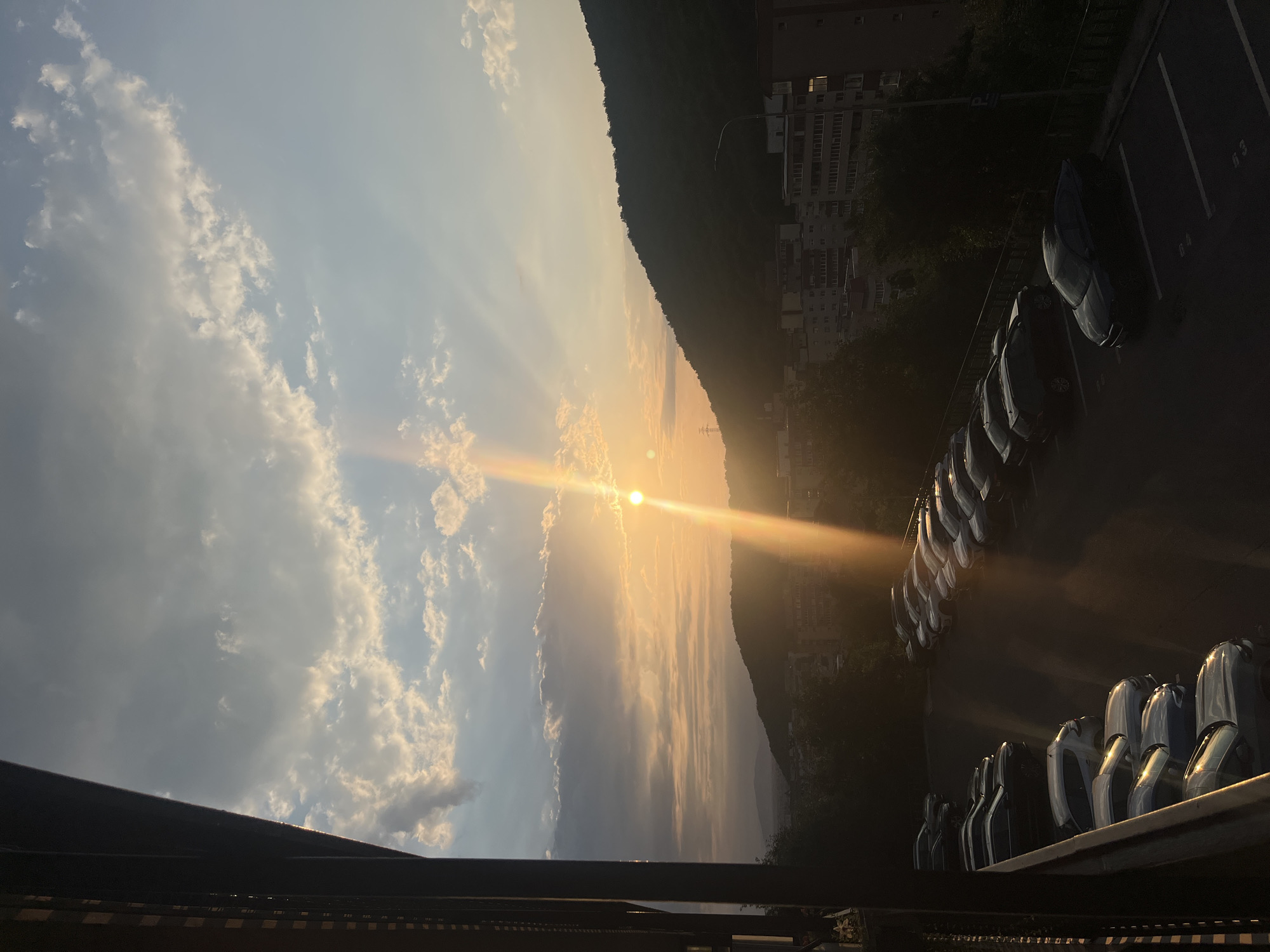 The image depicts a sunset scene over a residential area in Brasov, Romania, likely from a balcony or elevated viewpoint. The sun is low on the horizon, casting long rays of light through scattered clouds. A dark, silhouetted mountain range dominates the right side of the frame, with a few buildings visible at its base. In the foreground, a parking lot is filled with several cars, their surfaces reflecting the warm light of the sunset. The left side of the image features the dark, textured structure of a building or railing, creating a stark contrast with the illuminated sky and landscape. The overall mood is serene and atmospheric, characteristic of a late afternoon in a European city.