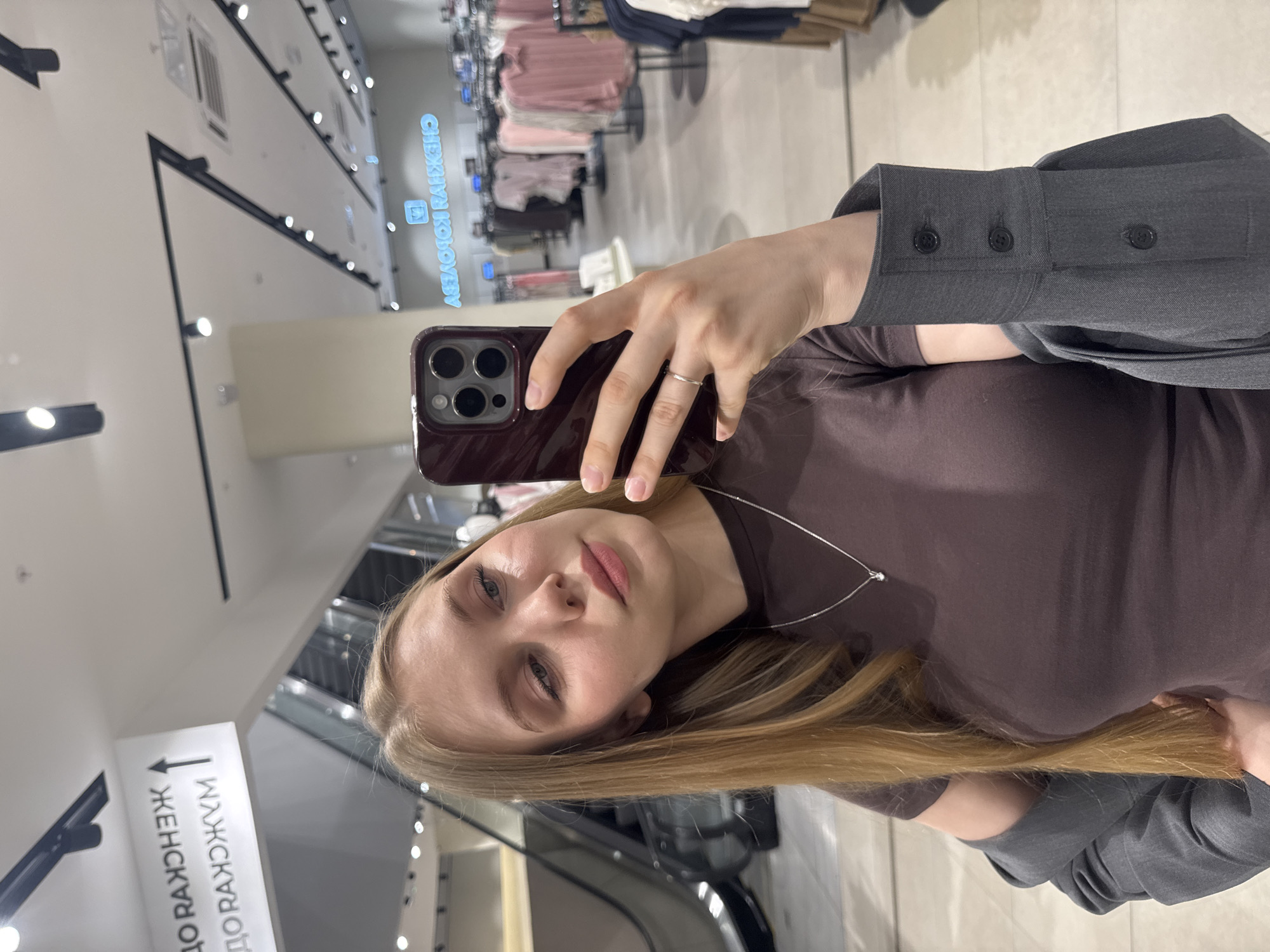 A young woman with long, light brown hair is captured in a mirror selfie inside a shopping mall or large department store in Perm, Russia. She is wearing a dark brown, short-sleeved top and a charcoal grey blazer, with a thin silver necklace featuring a small pendant. Her left hand holds a smartphone with a dark red case, its triple-lens camera module visible. She is looking at her reflection in the mirror.
In the background to her left, an escalator is visible, ascending to an upper floor. Above the escalator, a white directional sign, reflected in the mirror, displays Cyrillic text: "ЖЕНСКАЯ" (Women's) and "МУЖСКАЯ" (Men's) with an upward arrow, indicating these departments are on a higher level.
The background to her right, also reflected in the mirror, shows a clothing retail space. Racks of clothing, predominantly light-colored shirts and blouses, are arranged throughout the area. A prominent neon sign on the back wall, reflected in reverse, clearly reads "ЖЕНСКАЯ КОЛЛЕКЦИЯ" (Women's Collection). The environment is brightly lit by numerous track lights and recessed fixtures on a white ceiling, which also features visible air conditioning vents. The floor appears to be a light-colored tile or polished surface.