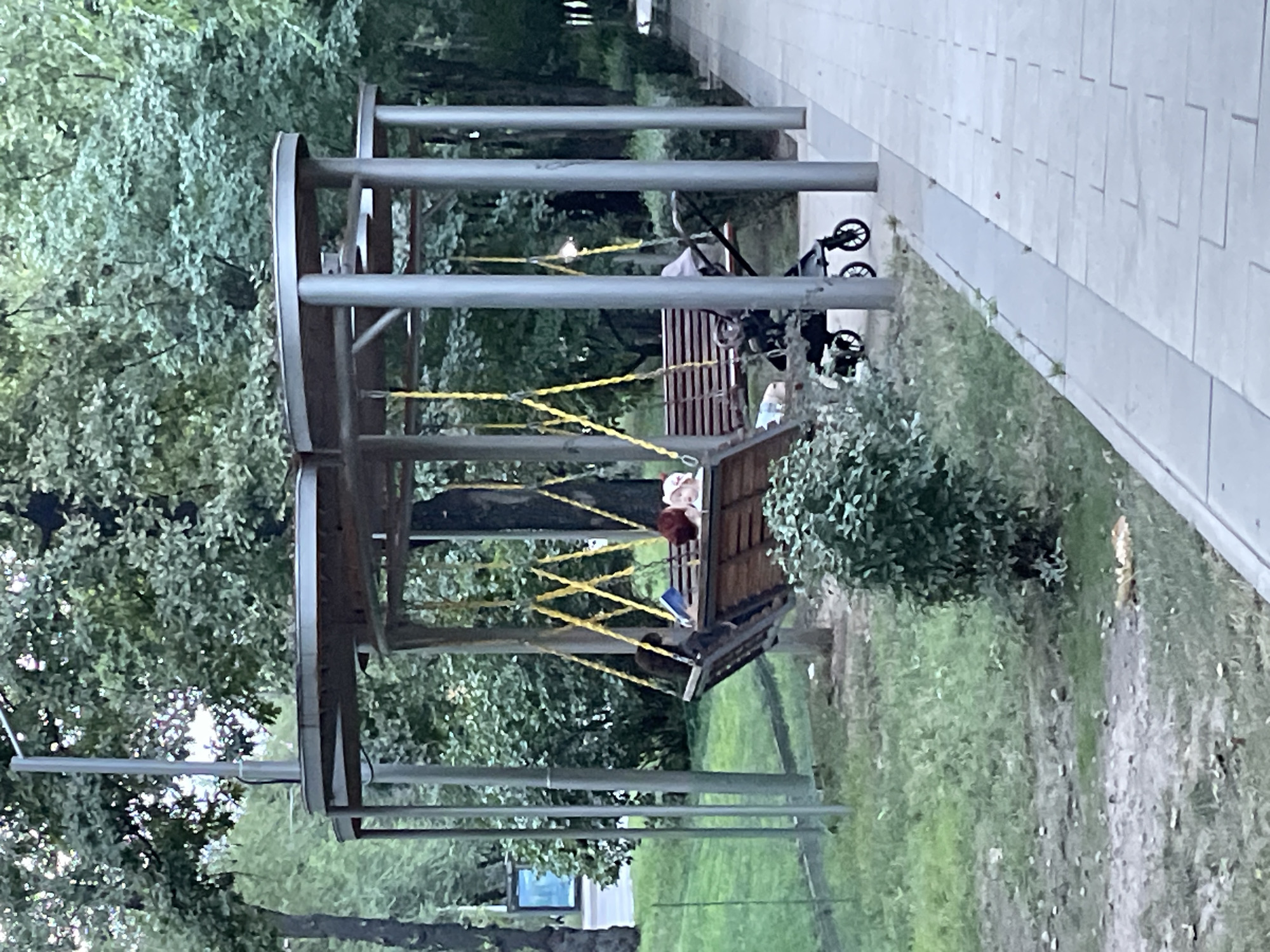 In Almaty, Kazakhstan, within a park-like setting, a rustic wooden swing set with a canopy structure is occupied by two individuals. One person, positioned towards the left of the swing, appears to be reading a book, indicated by the object held in their hands. To their right, another person is reclining with their head resting on a pillow. A baby stroller, dark in color, is situated to the right of the swing. The environment is characterized by lush green trees and foliage, with a paved walkway running parallel to the grassy area. The lighting suggests it is daytime, possibly overcast.