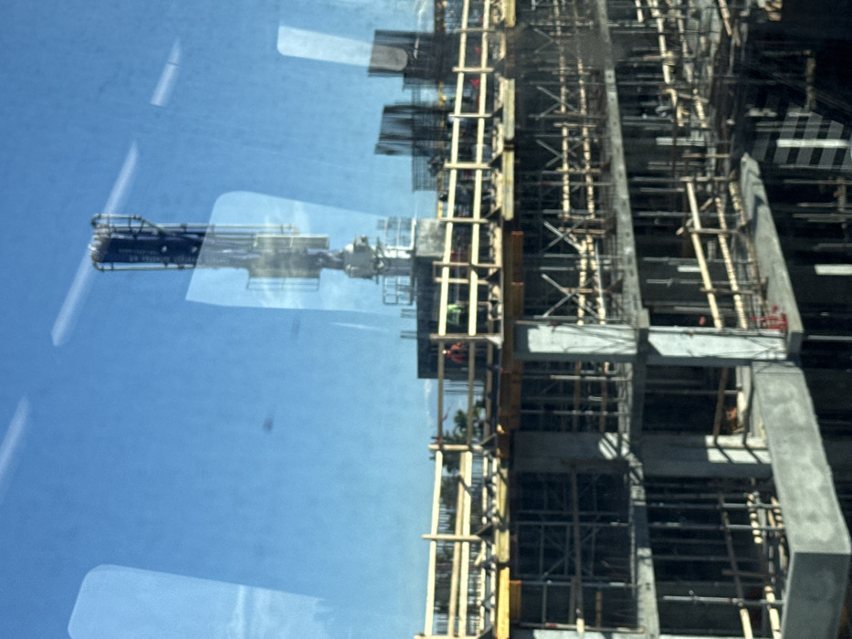 This image depicts an active multi-story building construction site in Hialeah, United States of America, viewed from an elevated perspective through a reflective glass pane. The primary subject is a concrete structural frame in various stages of completion, extending upwards through multiple levels. Extensive wooden formwork and scaffolding support the concrete slabs and columns on the visible floors. On the uppermost completed deck, several sections of vertical rebar cages are visible, indicating areas awaiting concrete pours. A large, articulated concrete pump boom extends vertically from a central point on the structure, its arm retracted. Three construction personnel are present on the top deck; one wears an orange high-visibility vest, and at least two others wear green or yellow high-visibility vests. They appear to be engaged in supervisory or operational tasks on the unfinished floor. The sky overhead is clear and bright blue. Distant palm trees are faintly visible on the horizon, consistent with the geographic location.