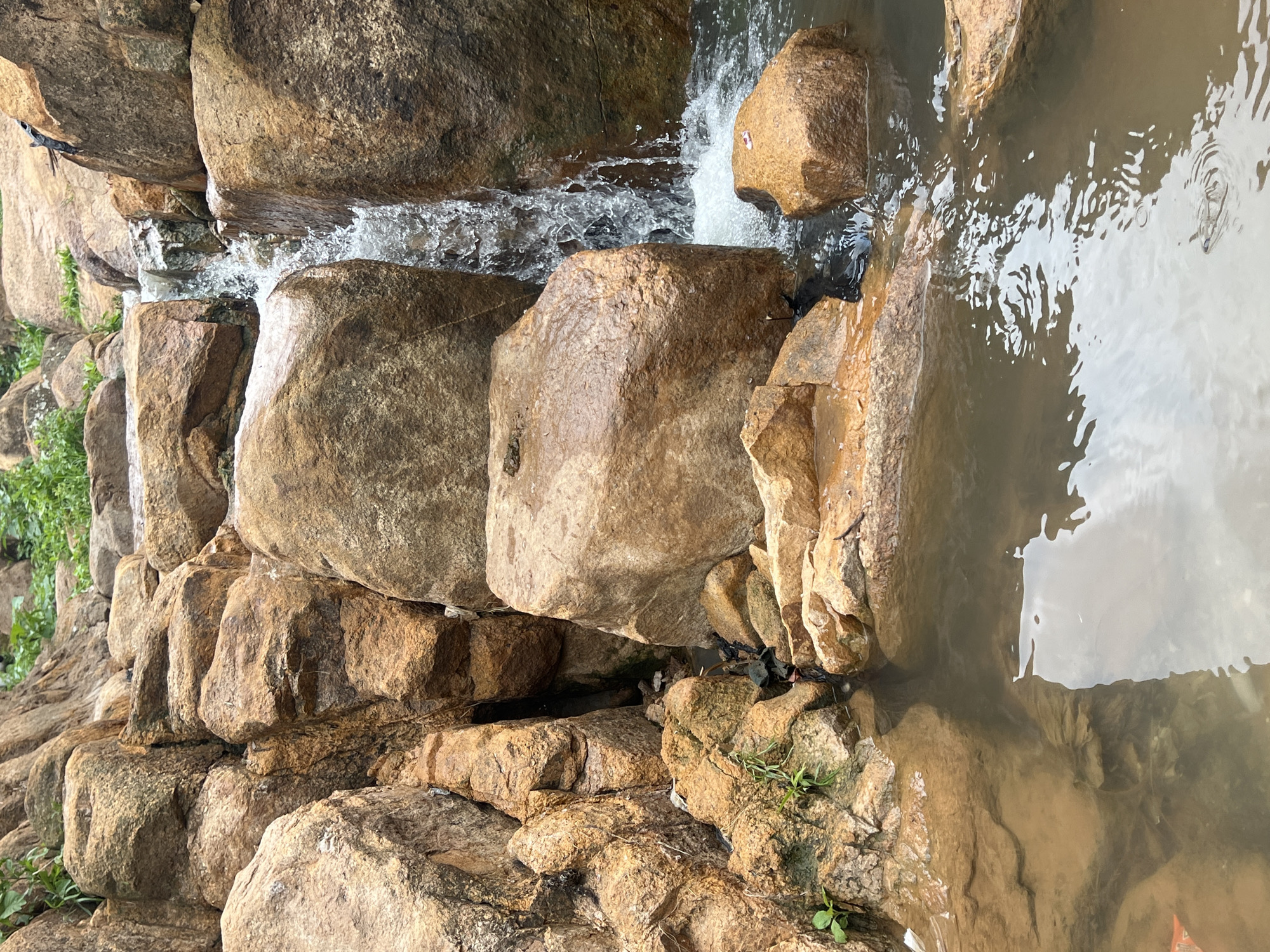 A close-up, eye-level shot captures a small, man-made waterfall cascading over rough-hewn, reddish-brown rocks. The water, appearing somewhat murky, forms a small pool at the base, reflecting the cloudy sky above. Green vegetation sprouts from crevices in the rocks, adding a touch of life to the scene. The overall impression is one of a naturalistic water feature, possibly within a park or garden setting.