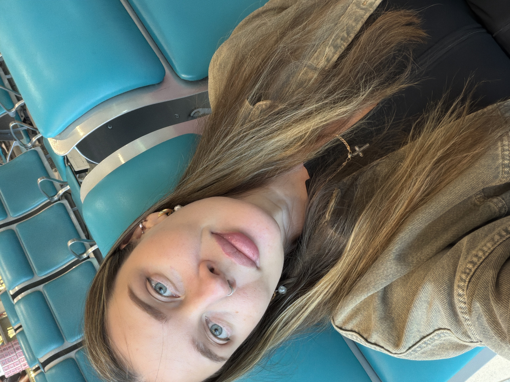 A young woman with fair skin, light brown hair with blonde highlights, and light blue eyes is in Domodedovo, Russia. She is wearing a nose ring, earrings, a cross necklace and a tan jacket. She is sitting on a row of turquoise airport seats.