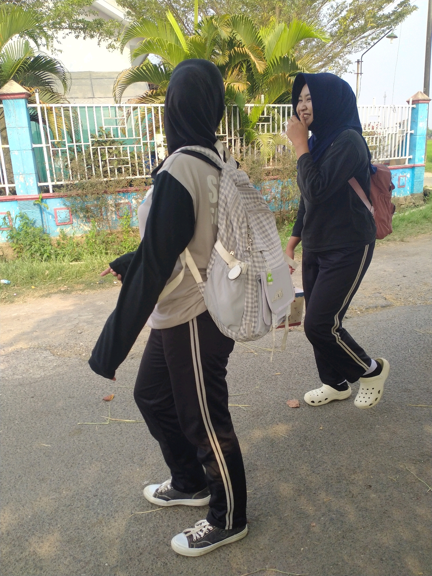 Two young females wearing hijabs are walking on a paved road in Begajah, Indonesia. The female on the left wears a grey t-shirt with black sleeves and black pants with white stripes, and carries a grey checkered backpack. The female on the right wears a black long-sleeved shirt, black pants with white stripes, and a maroon backpack. She is smiling and touching her face with her hand. They are walking past a blue and white fence with palm trees and green vegetation behind it. The road appears to be a rural area.