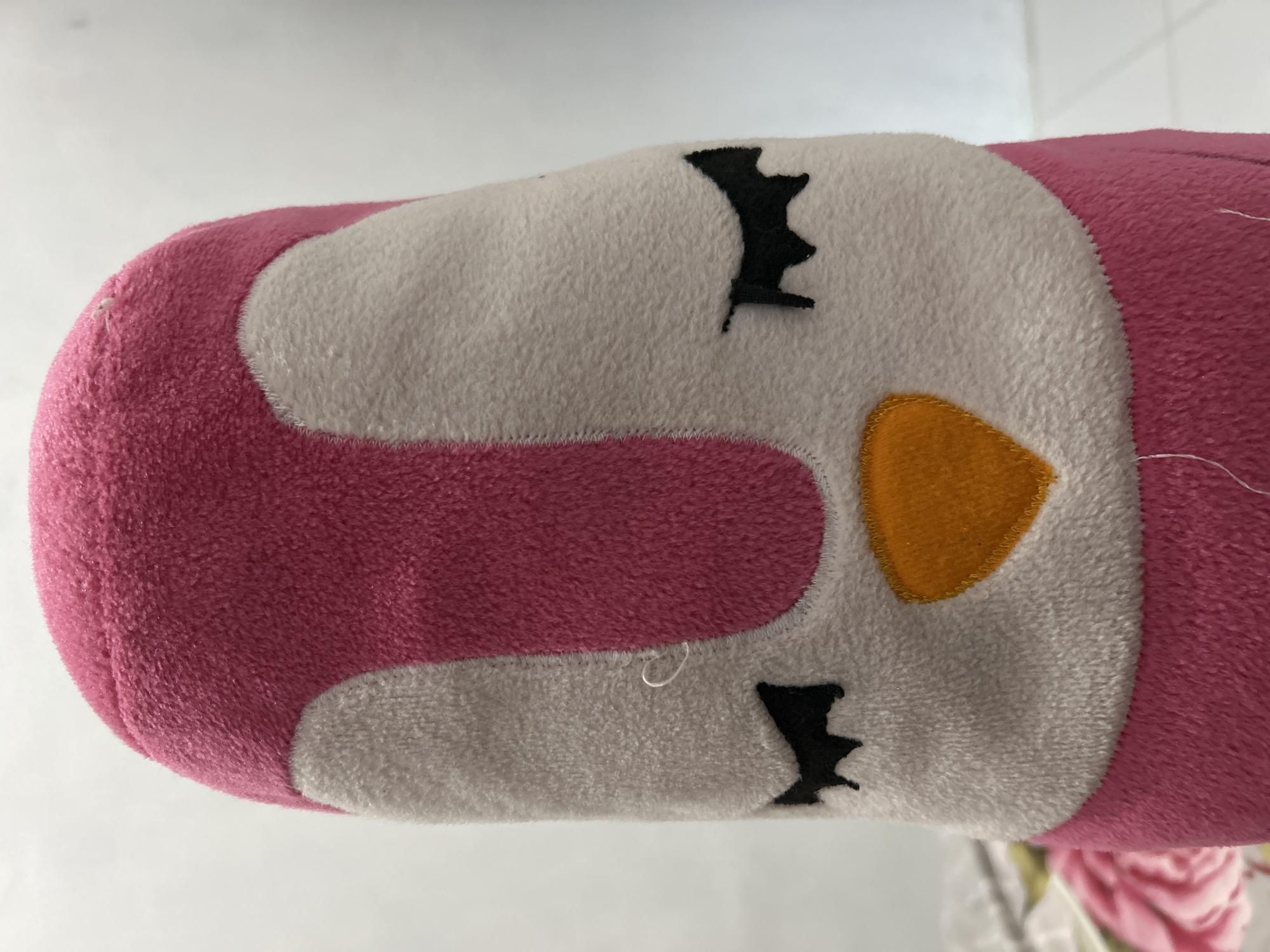 A close-up, slightly off-center image shows the head of a pink and white stuffed toy. The toy's head is cylindrical, with a pink outer layer and a white face. Black, stylized eyelashes are stitched onto the white face above and below a yellow beak. A white thread is visible near the lower eyelash. The background is a blurry white surface, possibly a table or countertop. The image does not provide any contextual information to suggest a location in Lhokseumawe, Indonesia, or any details about people, actions, interactions, or the environment beyond the immediate subject.