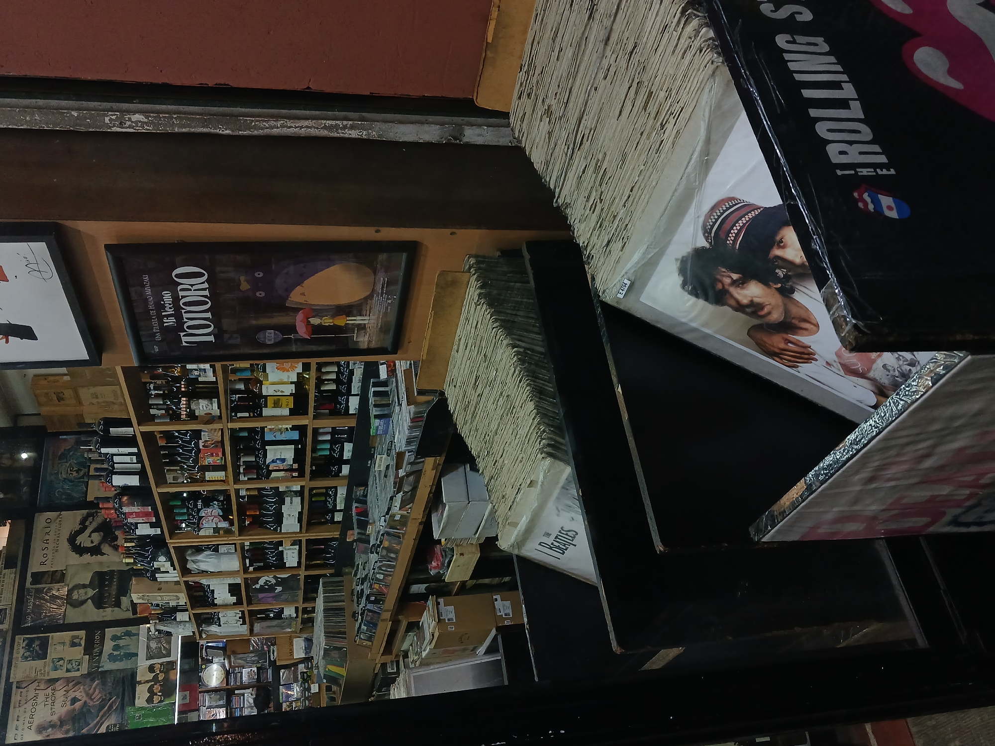 The image shows a view inside a record store in Buenos Aires, Argentina. Records are densely stacked on shelves and on top of each other. Prominent album covers visible include "The Rolling Stones," "The Beatles," and "Totoro." Bottles, likely wine, are arranged on shelves behind the records. Posters and advertisements are also visible on the wall, including one for Rosario, and for a movie called "Ventarrón," which mentions Cesaria Evora.