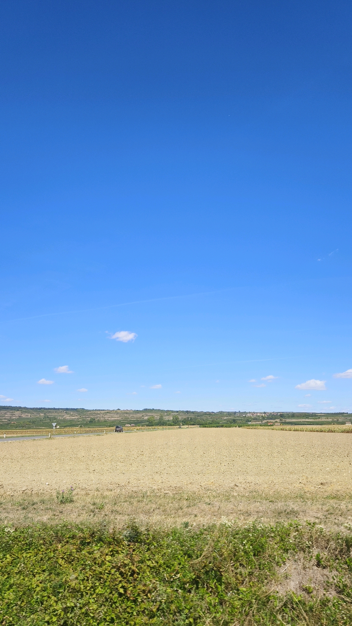 This image captures a rural landscape under a clear blue sky with sparse white clouds. In the foreground, there is a patch of green vegetation. Beyond this, a large, tilled field occupies the mid-ground, appearing a light tan color. A paved road runs horizontally across the scene, with a dark-colored car driving on it. Further in the distance, there are rolling hills with scattered trees and buildings, suggesting a small settlement or village. The overall impression is one of an agricultural area on a sunny day.