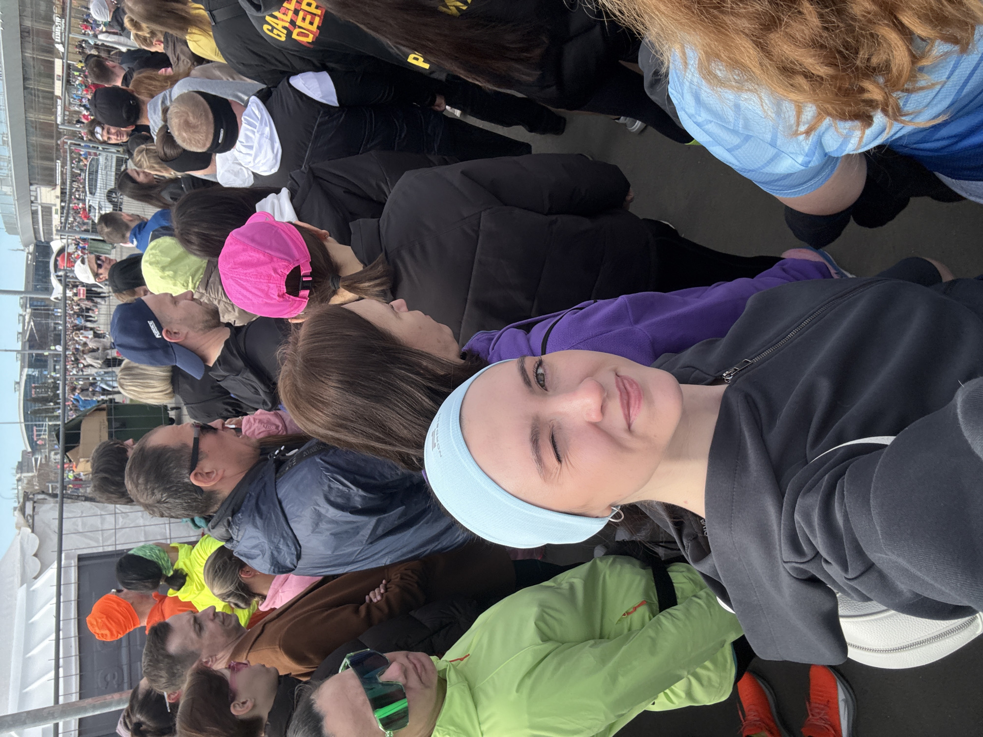 A selfie taken at eye-level shows a crowd of people outdoors. The photo's subject, a fair-skinned woman wearing a light blue headband and dark jacket, winks at the camera. To her left, a woman in a purple shirt looks away, and a man in sunglasses wears a bright green jacket. The crowd behind them consists of people with various hats, jackets, and hairstyles. In the background, a metal fence separates the crowd from a structure in the distance. "Kazan, Russia" is not discernible from the visual information provided.