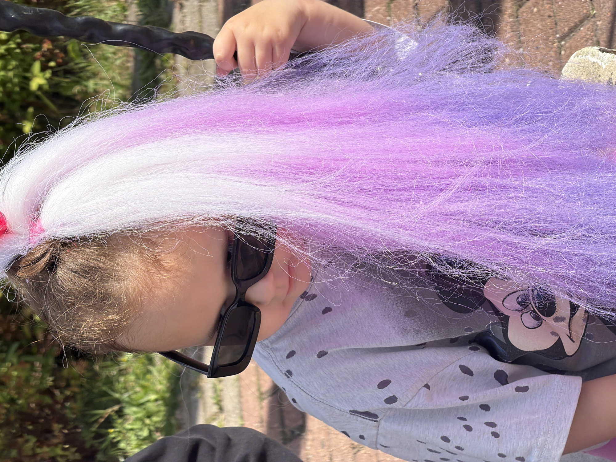 A close-up, medium shot captures a young person from the shoulders up, outdoors in daylight. The person is wearing black, rectangular sunglasses and a grey t-shirt with a scattered pattern of dark spots and a graphic of Mickey Mouse. Their hair is styled in a high ponytail, adorned with a pink accessory. The ponytail cascades down, featuring a blend of white and pastel purple artificial hair. The individual is holding onto a dark, textured object, possibly part of a playground swing. The background consists of blurred green foliage and a glimpse of reddish-brown pavement. There is no indication of the image being taken in Moscow, Russia.