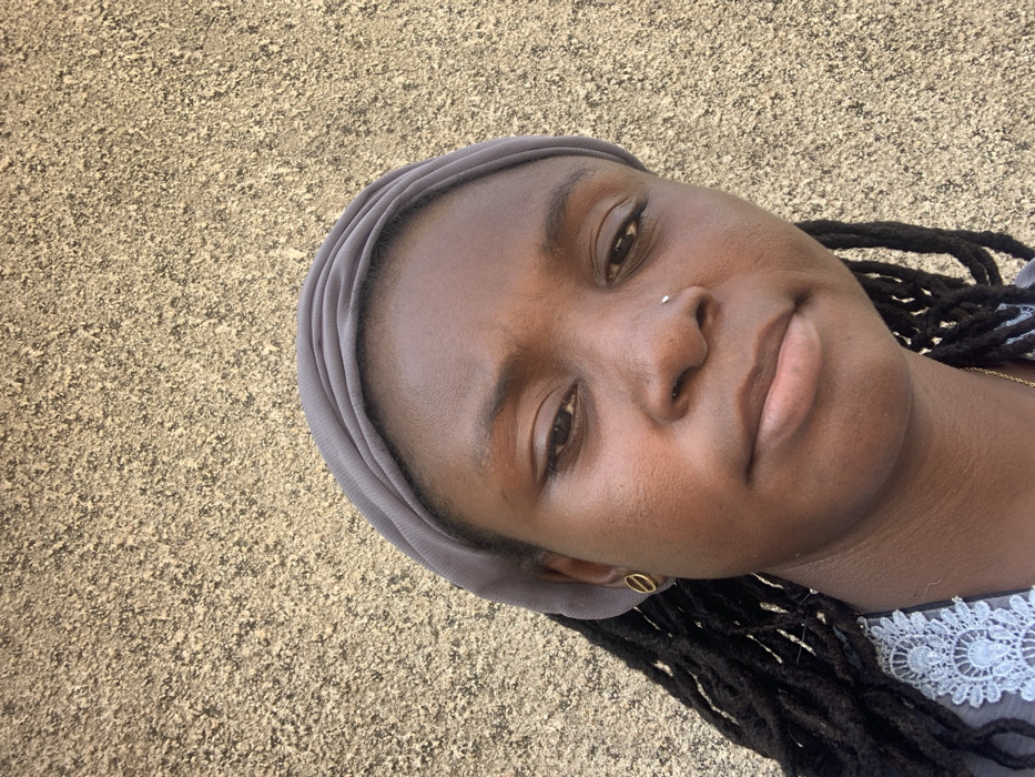 The image depicts a close-up portrait of an adult female, situated in Bojude, Nigeria, against a uniform, textured light-brown wall. The subject is positioned centrally, facing directly towards the camera with a neutral expression. She has dark skin, with visible dreadlocks or braids extending from beneath a grey fabric head covering. A small silver stud piercing is present on her left nostril, and a golden, oval-shaped stud earring is visible in her left earlobe. The neckline of a garment with light blue or grey lace detailing is partially visible on her upper chest, along with a thin golden chain. The background wall exhibits a consistent rough, granular texture. Lighting appears natural, casting subtle illumination on the subject's face and the wall.
