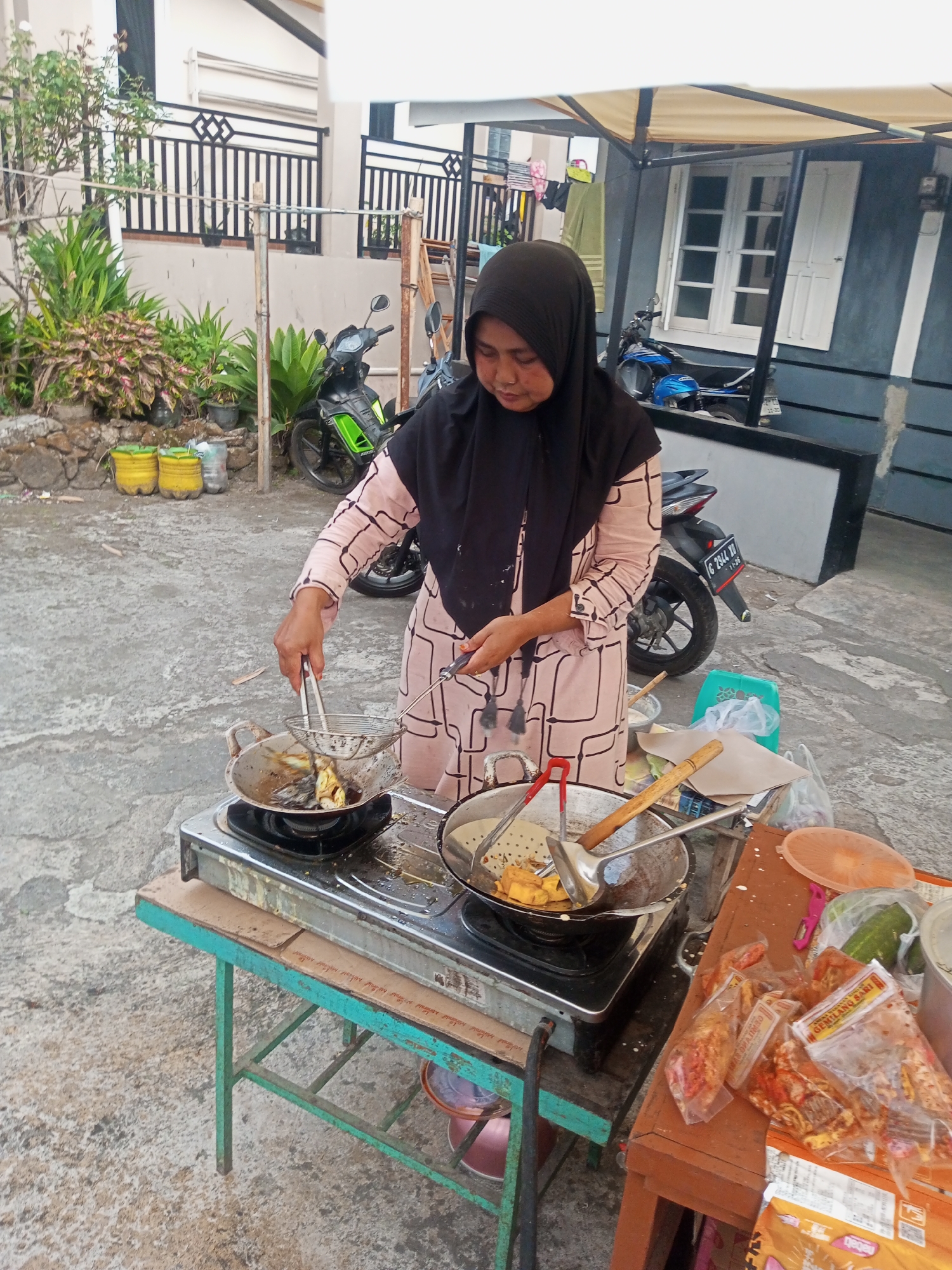 In Pandansari, Indonesia, a woman wearing a black hijab and a pink patterned dress is actively frying food on a portable gas stove. She is using tongs to handle items in a pan of hot oil, with a skimmer and ladle nearby. The stove sits on a green metal table. To the right, a wooden table holds various items, including packaged snacks, cucumbers, and containers. In the background, two motorcycles are parked, one a green and black model and the other a blue scooter. The scene appears to be outdoors, possibly a street food stall, with a building and a fence visible behind the woman and plants. The ground is paved with concrete.
