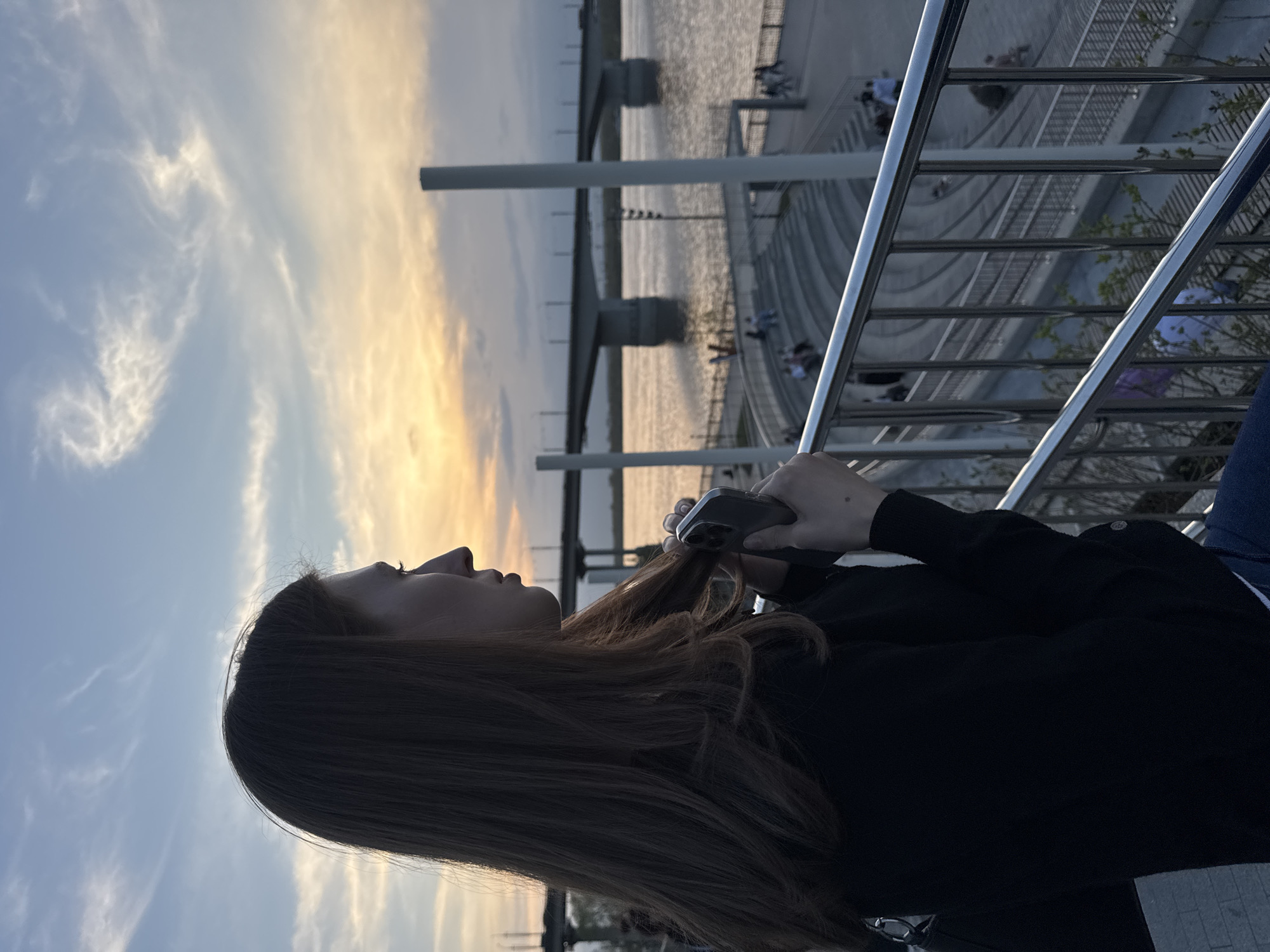 A medium shot depicts a woman holding a smartphone near a riverbank. She is standing by a metallic guardrail, with the river and a bridge in the background. In the distance, other people are visible. The sky is filled with clouds, suggesting either sunrise or sunset. The scene is likely located in Perm, Russia, based on the architectural style of the bridge and riverbank.