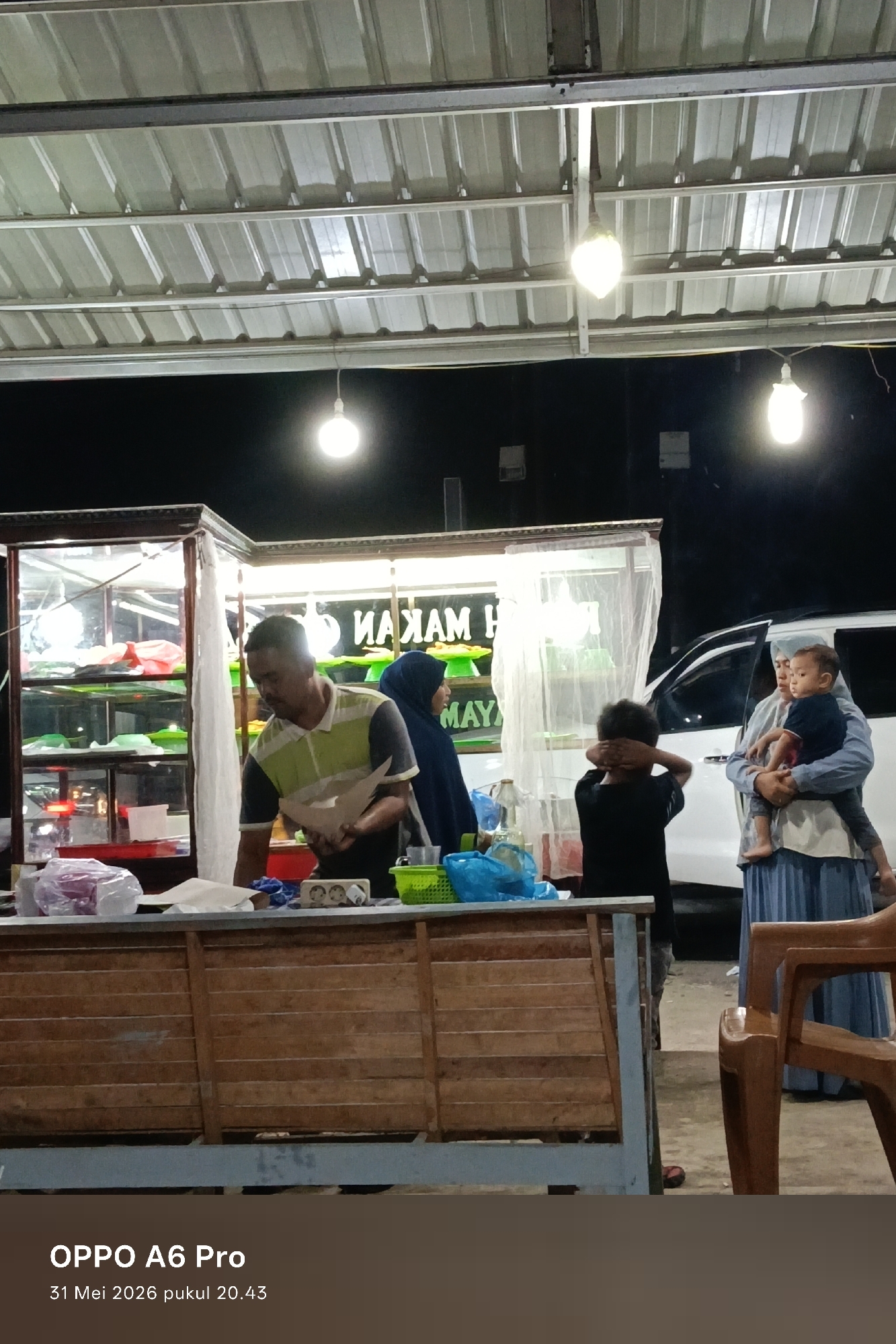 Here is a forensic description of the image:
The image shows a nighttime scene in what appears to be an outdoor food stall or small restaurant, possibly in Sangso, Indonesia. The timestamp indicates May 31, 2026, at 20:43.
In the foreground, a man wearing a striped polo shirt stands behind a wooden counter, appearing to prepare food or handle items. To the left, there's a glass display case containing food items.
To the right, there is a woman in a blue hijab and a woman in a long blue dress holding a child. A younger child stands in front of them with their hands covering their eyes. A white vehicle is visible in the background.
The area is lit by several hanging light bulbs under a corrugated metal roof. The ground appears to be unpaved. There is a stack of plastic chairs to the right. A plastic bin with blue contents is visible on the counter, along with a small green basket.