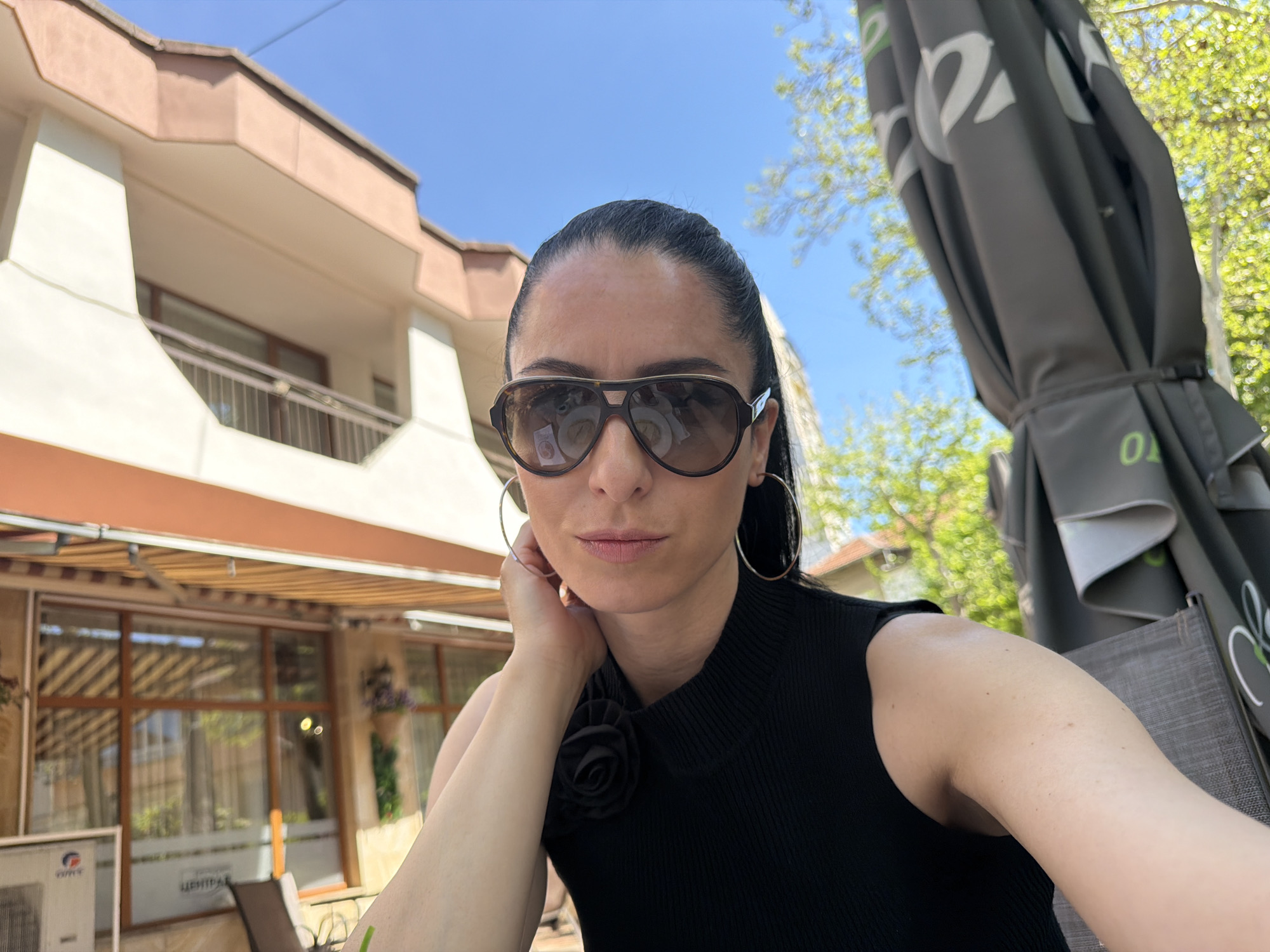 A woman takes a selfie outdoors, likely at a cafe or restaurant in Pleven, Bulgaria. She is the central subject, looking directly at the camera with a neutral expression. Her dark hair is pulled back, and she wears large, brown-framed aviator-style sunglasses that reflect the surroundings. She is adorned with large silver hoop earrings and a black, ribbed sleeveless top featuring a black fabric flower detail on her left shoulder. Her right hand is gently touching her jawline.
The scene unfolds on a bright, sunny day with a clear blue sky. To the woman's left, a multi-story building with a light-colored (white and pale brown) facade and balconies with metal railings is visible. A striped beige and brown awning extends over a lower section of the building, below which large windows reflect the environment. Inside one window, a white sign displays the Cyrillic text "ЦЕНТРАЛ" (Central), and an outdoor air conditioning unit clearly shows the brand "GREE."
To her right, a large, dark grey outdoor umbrella, partially folded and bearing white and green branding, is prominent. Behind the umbrella and the building, vibrant green foliage from trees indicates a lush outdoor setting. A portion of a dark grey woven chair is visible in the lower right foreground, suggesting outdoor seating. The overall impression is one of a relaxed moment in a sunlit urban outdoor space.