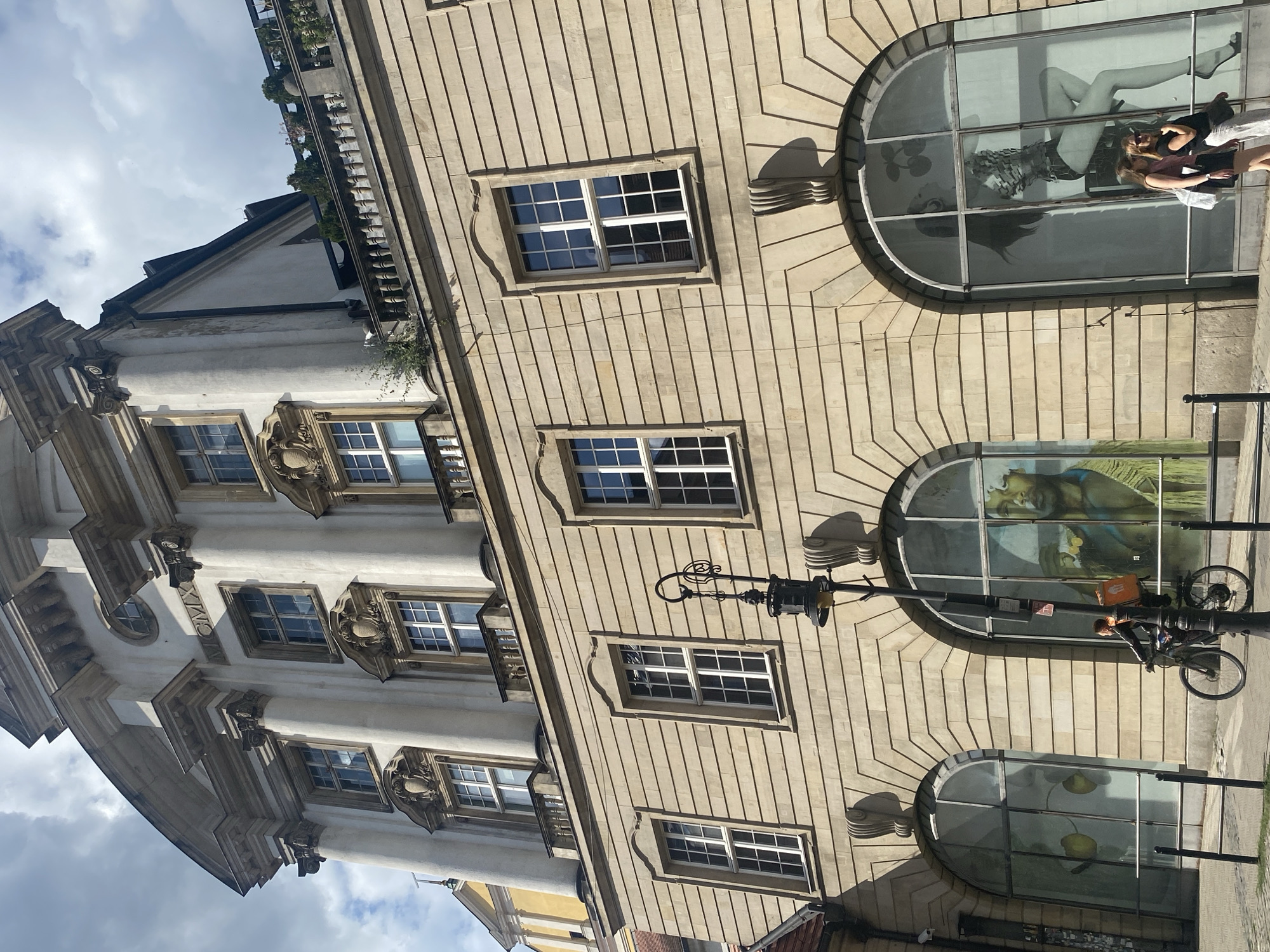 The image captures a street view in Poznan, Poland, featuring a large, ornate building with multiple stories and arched ground-floor windows displaying large artwork. The building's facade is constructed of light-colored stone with horizontal banding and decorative architectural elements, including pilasters, cornices, and elaborate window surrounds. Several windows are visible on the upper floors, with white frames and multiple panes. The uppermost section of the building has a curved roofline and classical detailing, including columns and pediments.
In the foreground, a person is riding a bicycle with an orange delivery bag on the back, traveling from left to right. They are wearing dark clothing. To the right of the cyclist, two women are standing on the sidewalk, talking and looking towards the building. One woman wears a dark skirt and a light-colored top, while the other has sunglasses and is wearing white pants and a dark top.
The ground-floor windows are particularly striking. The leftmost arched window appears to display a graphic with two yellow, lemon-like shapes. The central window features a large, vibrant mural of a person's face and upper body, rendered in shades of green, yellow, and brown, with a portrait-style presentation. The rightmost arched window showcases a large black and white graphic of a woman in a stylized pose, with what appears to be cherries above her.
A vintage-style black lamppost stands in front of the central and rightmost windows, adding to the urban streetscape. The sky above is partly cloudy, with patches of blue sky visible. The street pavement is visible in the foreground. The building's architecture suggests a historical or neoclassical style.