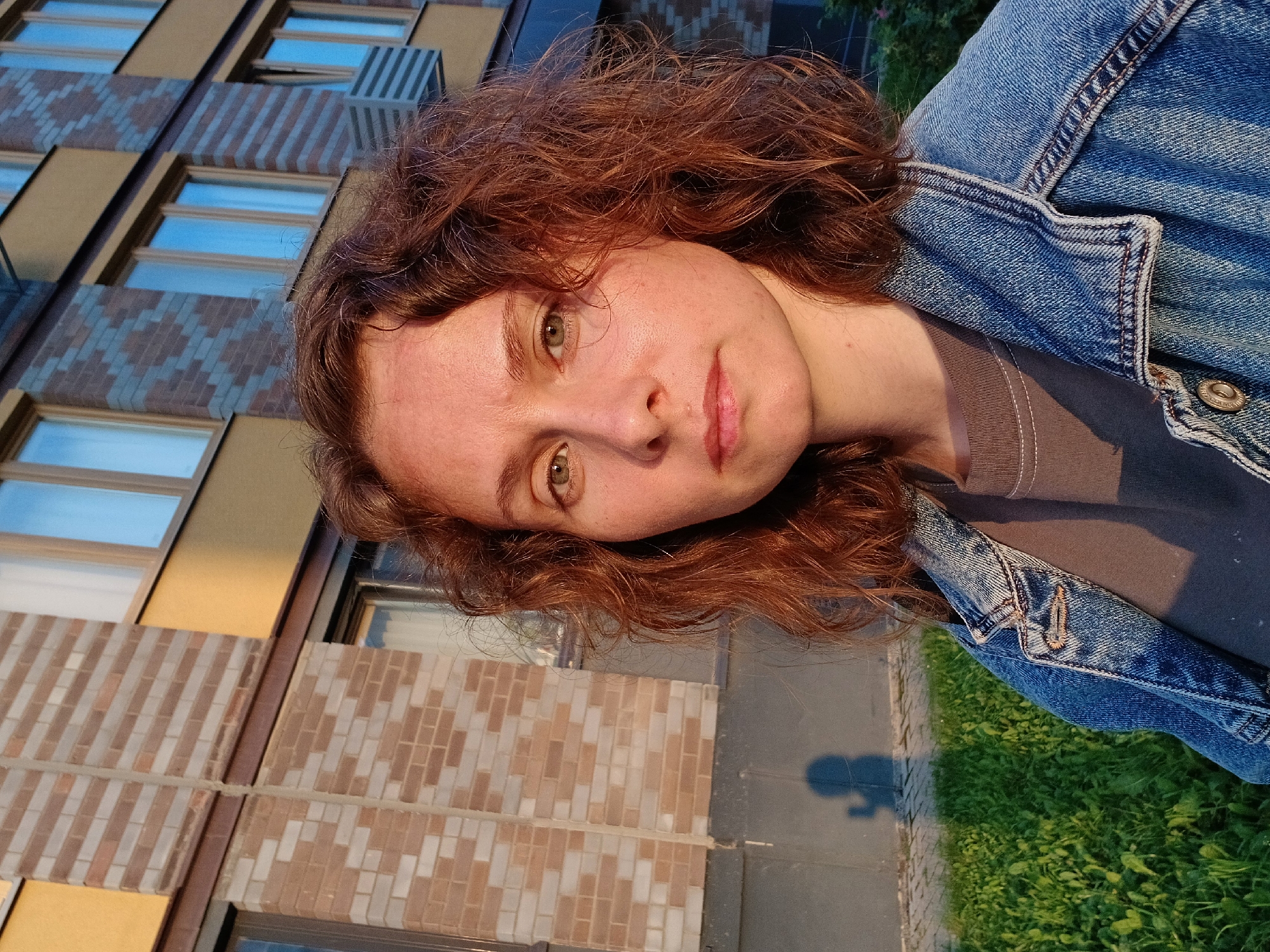 A medium close-up selfie captures a young woman with auburn, curly hair standing outdoors in front of a modern apartment building. The woman is facing the camera directly with a neutral expression, her eyes are green, and her skin has a slight flush from the sunlight. She is wearing a denim jacket over a brown t-shirt.
The background features a building with a facade of light brown and grey brickwork, accented with yellow panels. Several windows with reflective blue panes are visible. The lower part of the building has a darker grey base. To the left of the woman, a green grassy area is visible, with a shadow of a person holding a phone in their hand cast on the paved ground. The lighting suggests it is late afternoon or early evening, with warm sunlight hitting the scene.
The overall impression is a candid portrait set against an urban residential backdrop. Without specific landmarks or identifying features in the image, it is impossible to ascertain if the location is in Yekaterinburg, Russia.