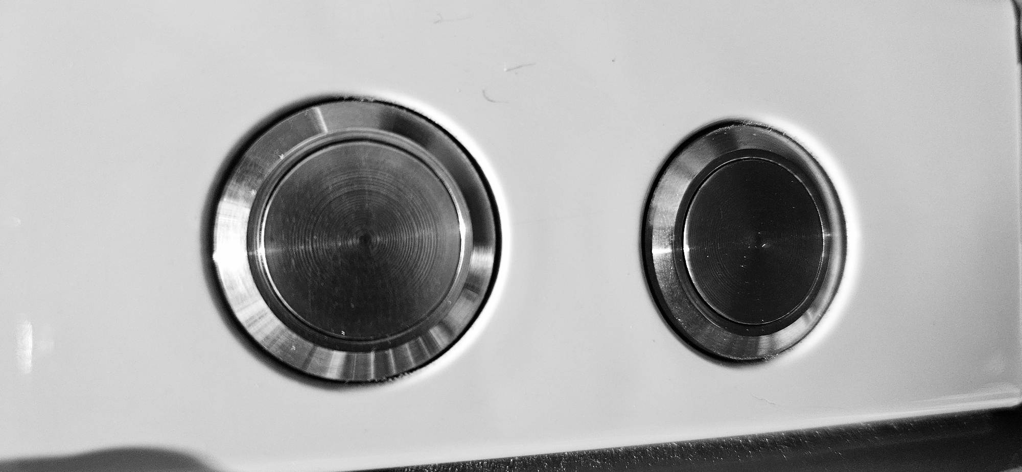 The image shows a close-up of two metal buttons set into a white surface. The buttons appear to be circular and flush-mounted. The left button has a concentric circular pattern on its surface, while the right button has a smoother, darker surface. The buttons are surrounded by a thin metal ring. The surface they are mounted on is white and slightly reflective. The image is in black and white. The location in the city of Plovdiv, Bulgaria is not discernible from the visual information.