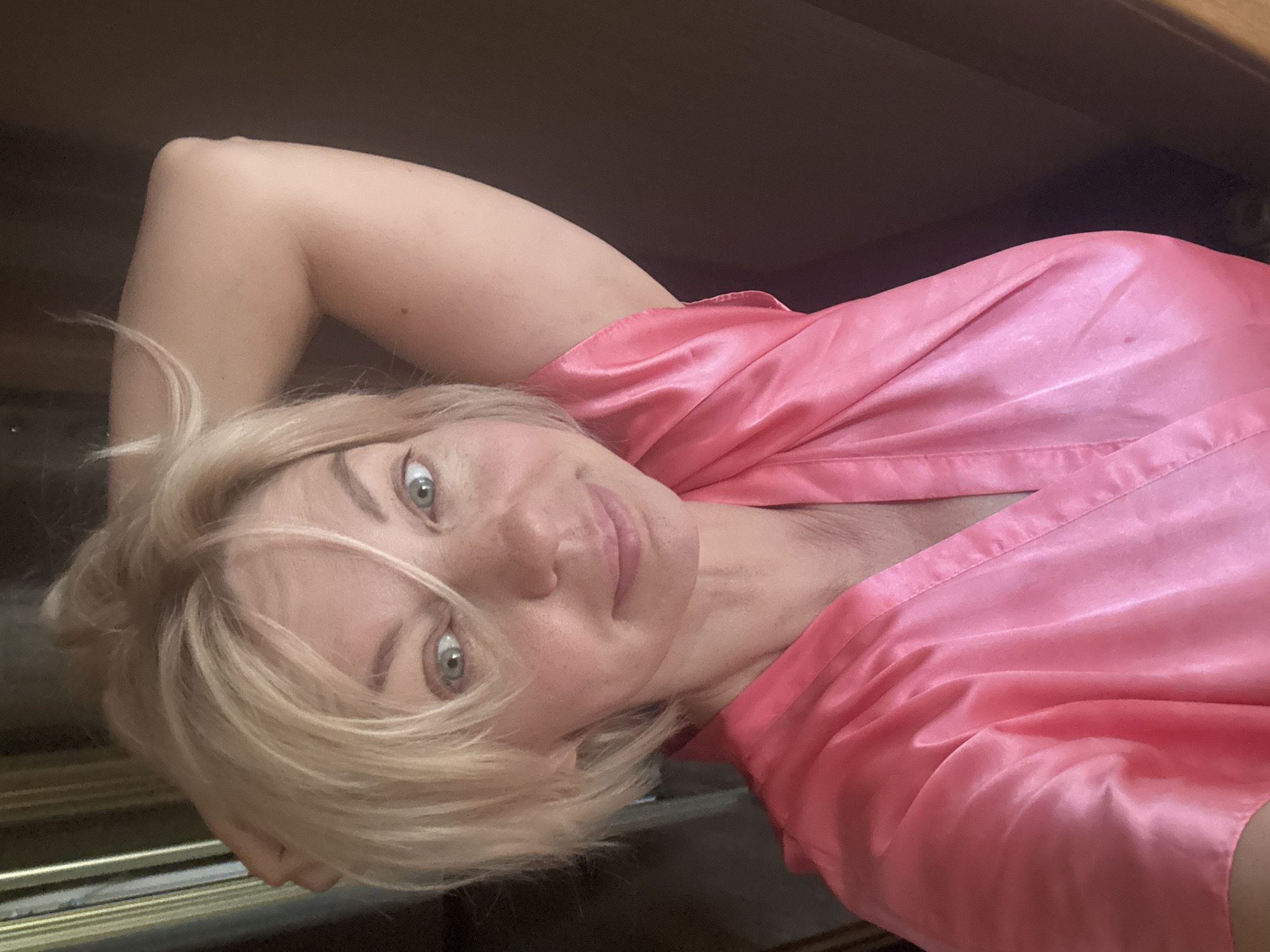 A medium close-up, selfie-style shot shows a Caucasian woman with short, blonde hair and blue eyes. She is wearing a pink satin robe and has her right arm raised, with her hand behind her head. Her expression is neutral, with a slight smile. The background is dimly lit and appears to be indoors, with a dark brown wall on the right side of the frame and a lighter, textured surface on the left. There is no information to indicate the location is Sochi, Russia.