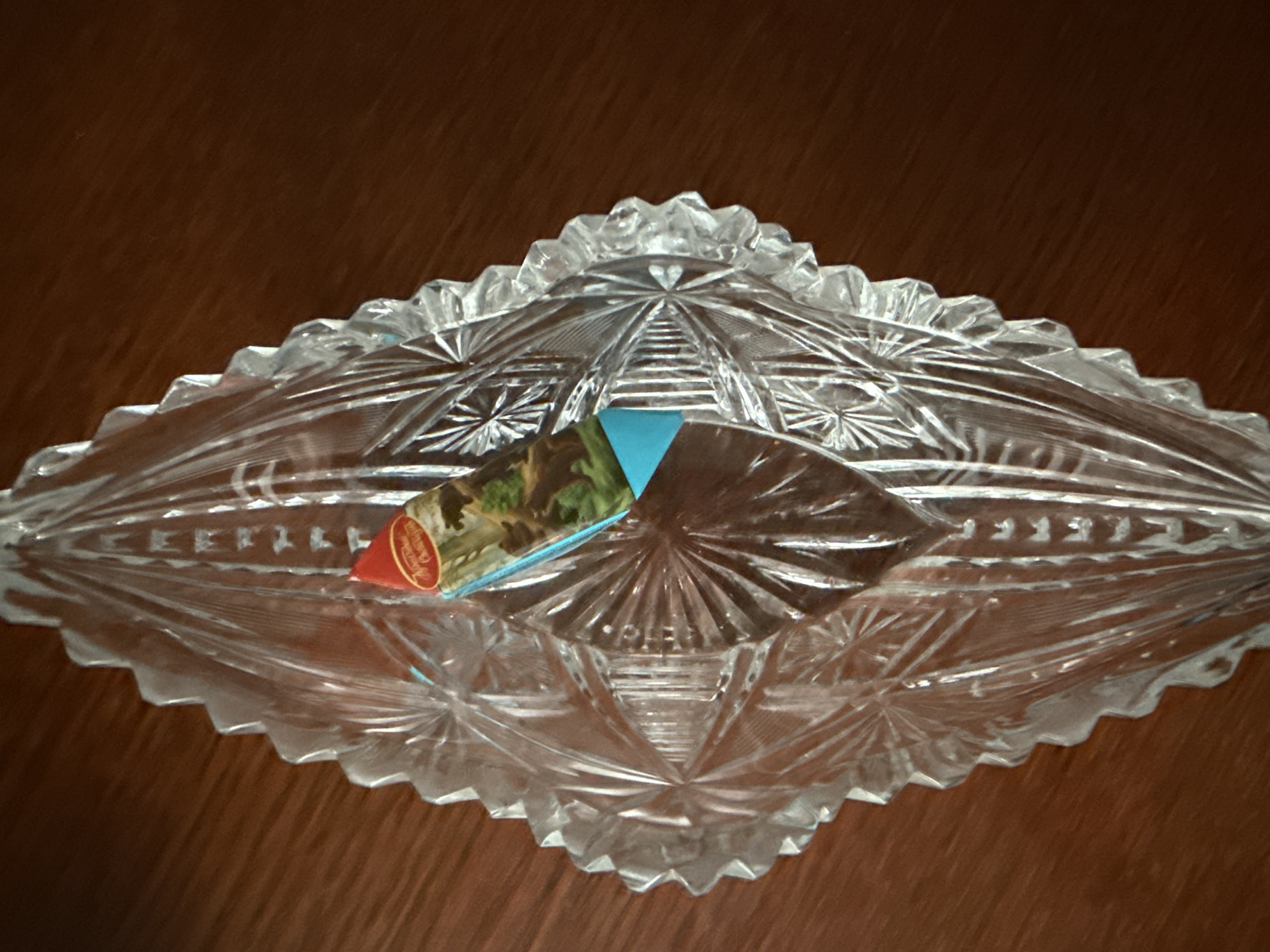 This is a close-up, overhead shot of a clear, cut-glass oval dish with a diamond pattern. The dish is resting on a dark brown, wooden surface, likely a table. The glass has many facets that refract light, creating a sparkling effect. Inside the dish, towards the center, there is a small, colorful label with a red circular border. The label appears to have some text in gold and a picture of a landscape with green trees and possibly a brown building. A triangular section of a blue material is visible beneath the label. The overall impression is one of a decorative item, possibly an antique or a piece of fine tableware. There are no people or animals visible. The lighting appears to be artificial, with some glare and reflections on the glass. The scene is static and lacks any discernible activity. The visible text on the label is not legible enough to be transcribed.