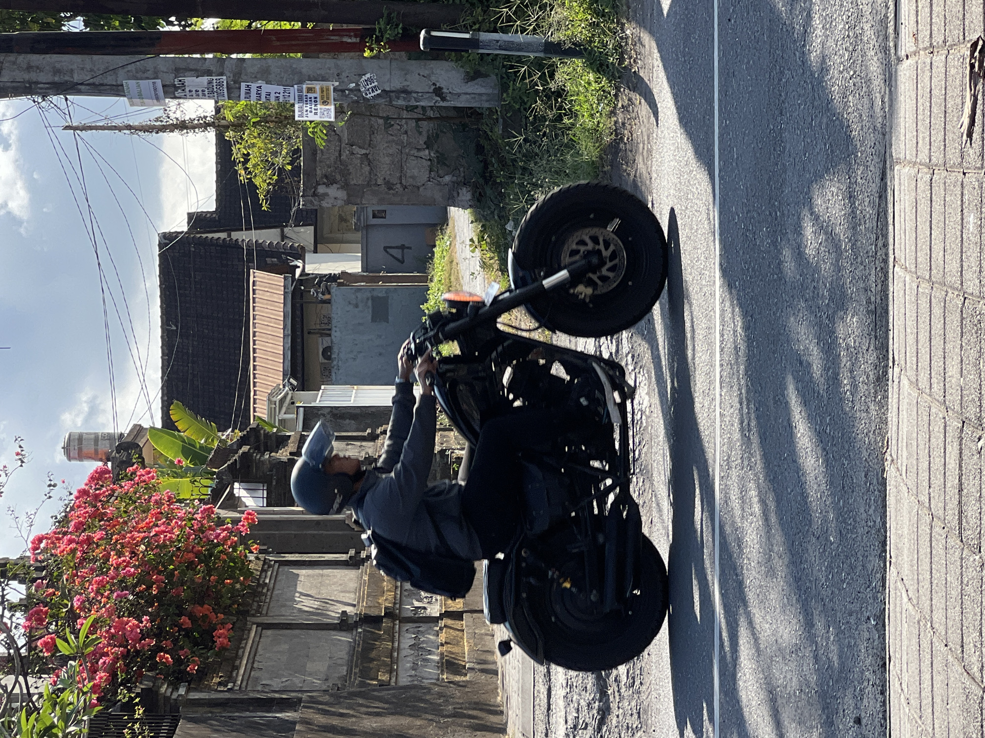 A person wearing a black helmet and a dark gray long-sleeved shirt rides a black custom motorcycle on a paved street. The person has a black backpack on. The motorcycle has a large front tire and a wide back tire. The scene is outdoors, with a building and a wall on the left side of the image. A tree with pink flowers is growing over the wall. On the right side of the image, there is a utility pole with several papers attached to it, and a small tree with green leaves. The background shows more buildings and vegetation. The sky is visible and appears to be partly cloudy. The sunlight casts shadows on the street, indicating it is daytime. The overall impression is a street scene in a residential area of Denpasar, Indonesia, given the style of the buildings, vegetation, and the presence of local signage.