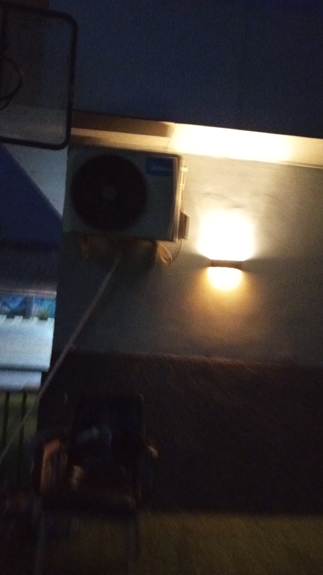 An exterior shot at night in Adankolo, Nigeria shows an air conditioning unit mounted on a wall. A wall-mounted light fixture illuminates the wall above a dark sofa and lawn. A blurry chair sits in front of the sofa. A piece of unidentified equipment appears to the left of the air conditioning unit.
