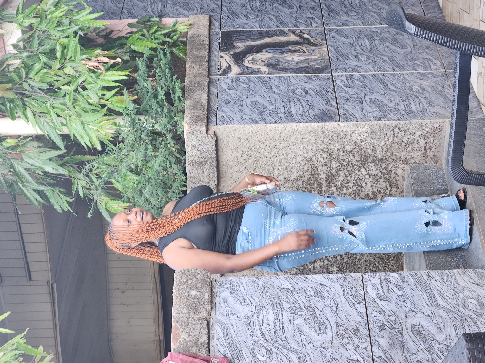 A full-length shot captures a woman wearing a black tank top and light blue, bedazzled, ripped jeans. She is lying on a set of exterior stairs of a building. The stairs are made of gray stone with visible seams, and they are partially adorned with green foliage and a black metal railing. The setting appears to be in an urban environment, Unknown, Unknown. The photograph is rotated clockwise approximately 90 degrees.