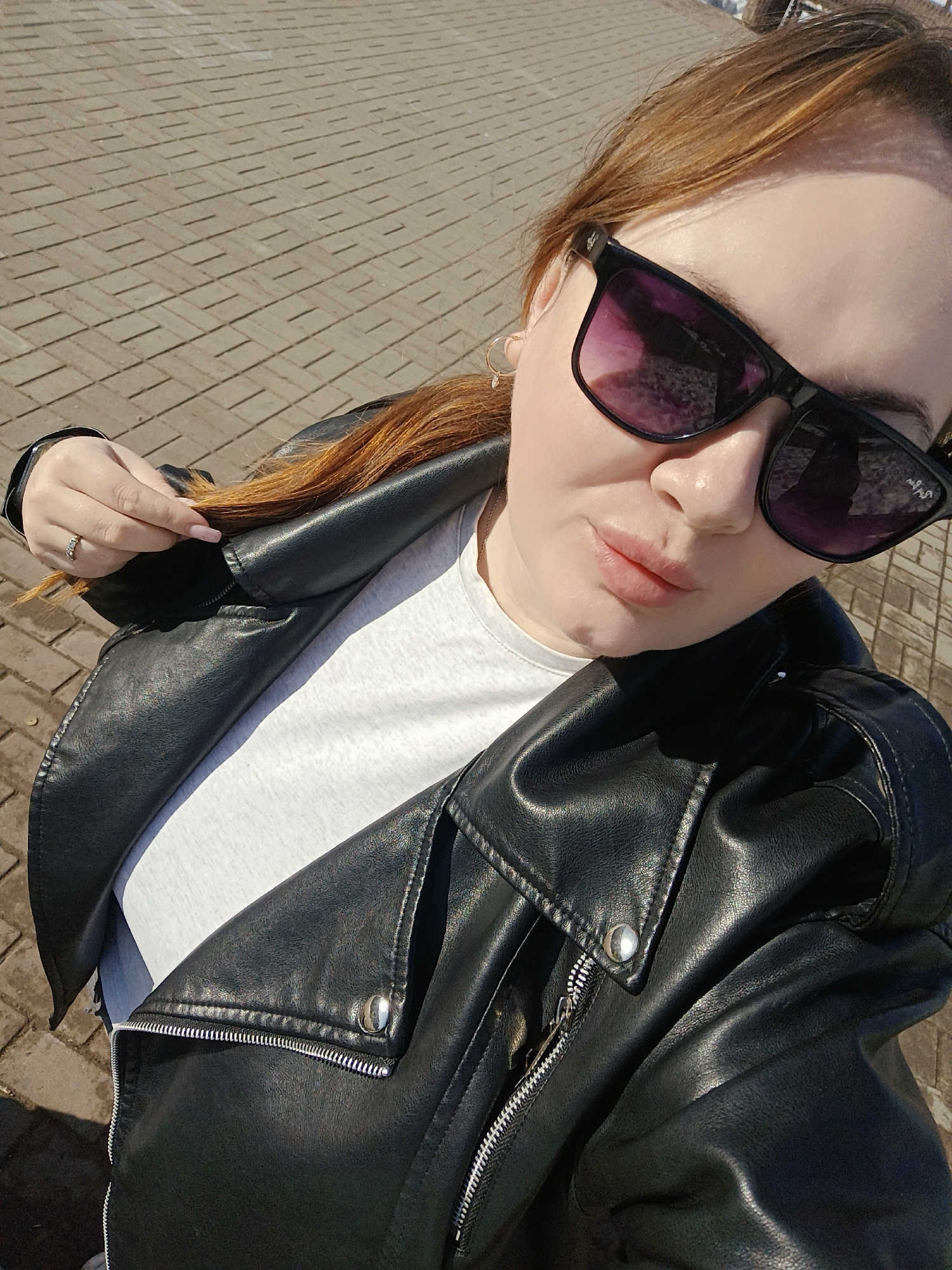 A woman with red hair, wearing sunglasses and a black leather jacket over a white t-shirt, is posing for a selfie outdoors. She has a ring on her finger and is holding a strand of her hair. The background shows a paved area with brick-like patterns. Based on visual details and the provided location, the scene is in Chaikovsky, Russia, although specific location details are unclear from the image itself.