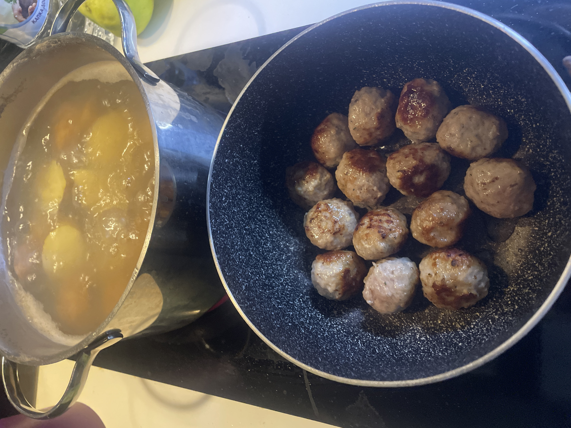 The image displays a cooking scene in a kitchen located in Sofia, Bulgaria. On the left, a silver-colored metal pot with handles contains a light yellow-brown liquid actively simmering or boiling, with several rounded objects, possibly potatoes or other vegetables, submerged. Adjacent to it on the right, a dark gray non-stick frying pan, characterized by a speckled surface, holds approximately fifteen to twenty cooked meatballs, exhibiting golden-brown and slightly seared exteriors.
Both the pot and pan are situated on a black cooktop, which appears to be ceramic or induction, with a white countertop visible to the left. In the background, behind the boiling pot, a green and yellow fruit (likely a pear or apple) is partially visible, along with a white and blue package. Cyrillic text is discernable on the package, reading "КАПКА.ДОБ...", with "КАПКА" meaning "drop" in Bulgarian, supporting the location context. A hand is minimally visible in the bottom-left corner, appearing to steady or hold the pot's handle. The scene captures the active preparation of a meal, with ingredients being boiled and meatballs being fried. The overall lighting is bright and even, consistent with an indoor setting.